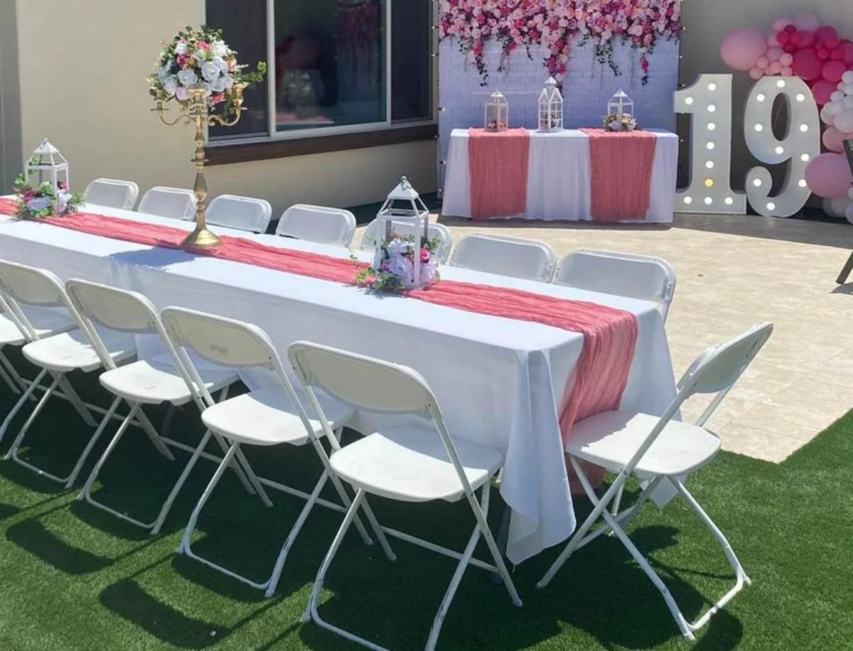 Outdoor party setup with a long white table decorated with pink table runners and floral centerpieces, white chairs, and a backdrop with pink flower decorations and large illuminated number 19, indicating a celebration or birthday party.