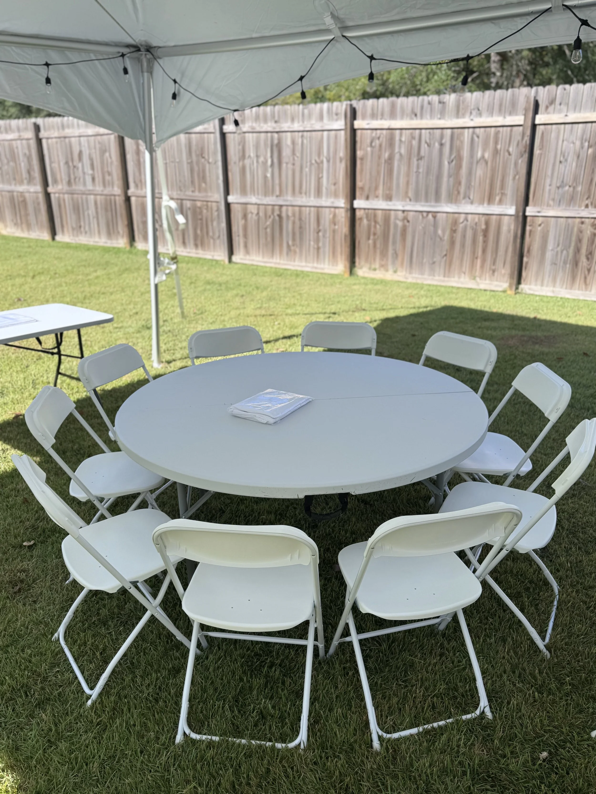 Outdoor gathering setup with a round white table and ten white folding chairs under a white canopy tent, with string lights overhead and a wooden fence in the background.