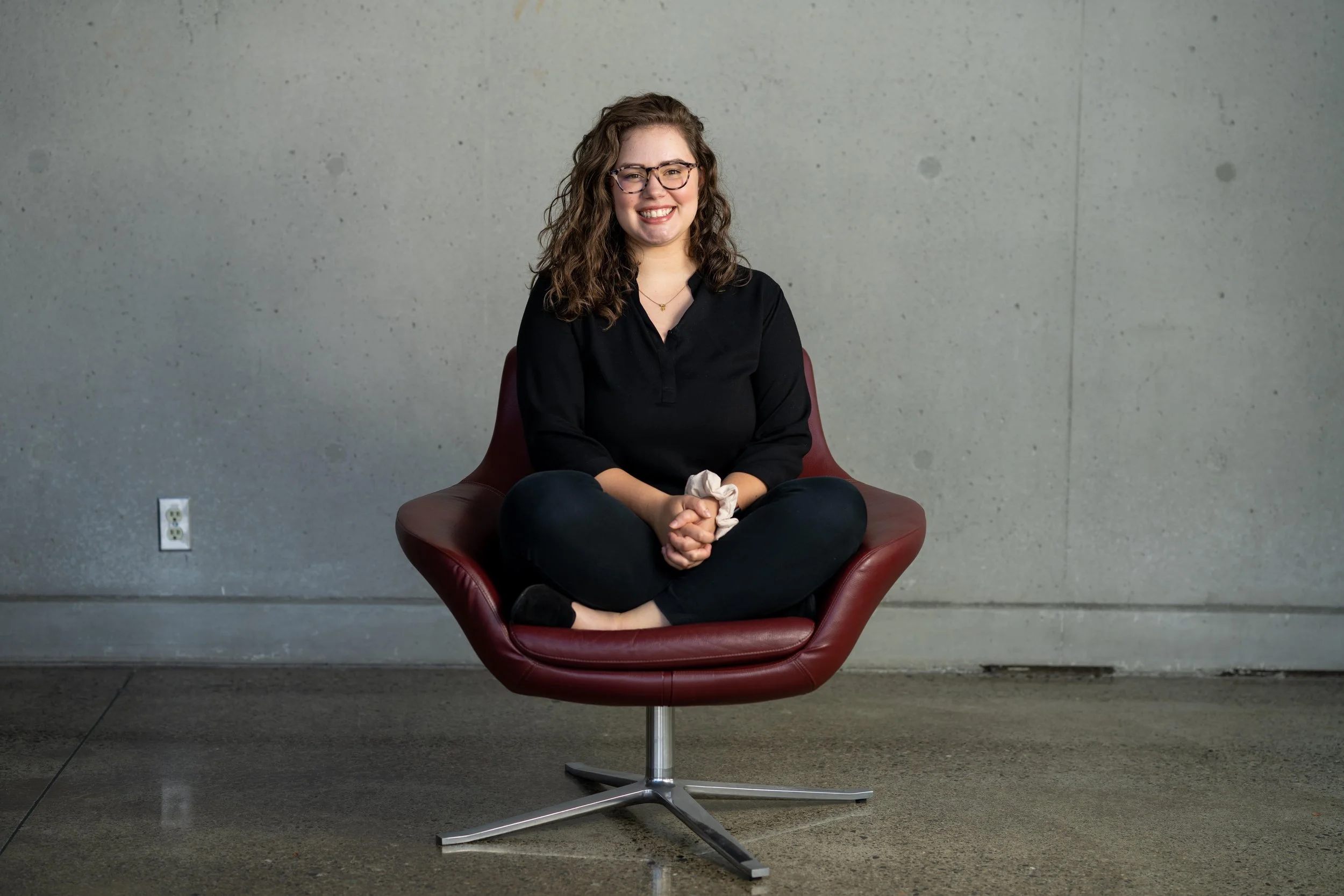 A woman with curly brown hair, glasses, and a black shirt sitting cross-legged on a modern red chair, smiling in front of a plain gray wall.