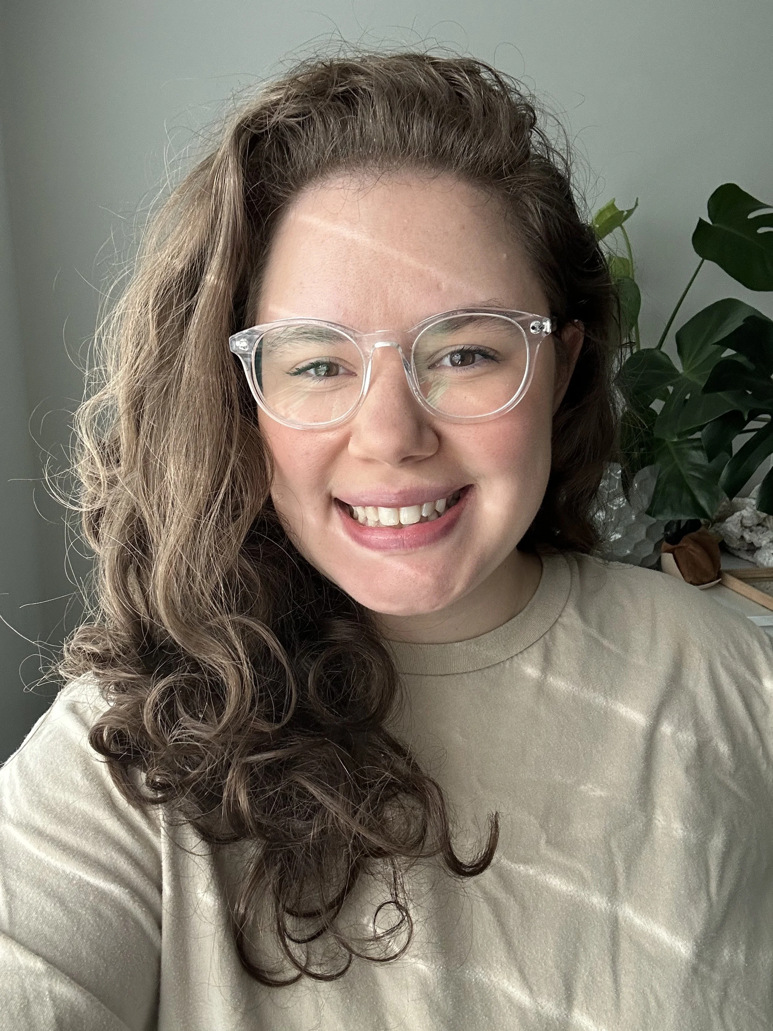 A woman with curly brown hair, wearing glasses and a beige shirt, is smiling at the camera. There is a green plant in the background on the right side.