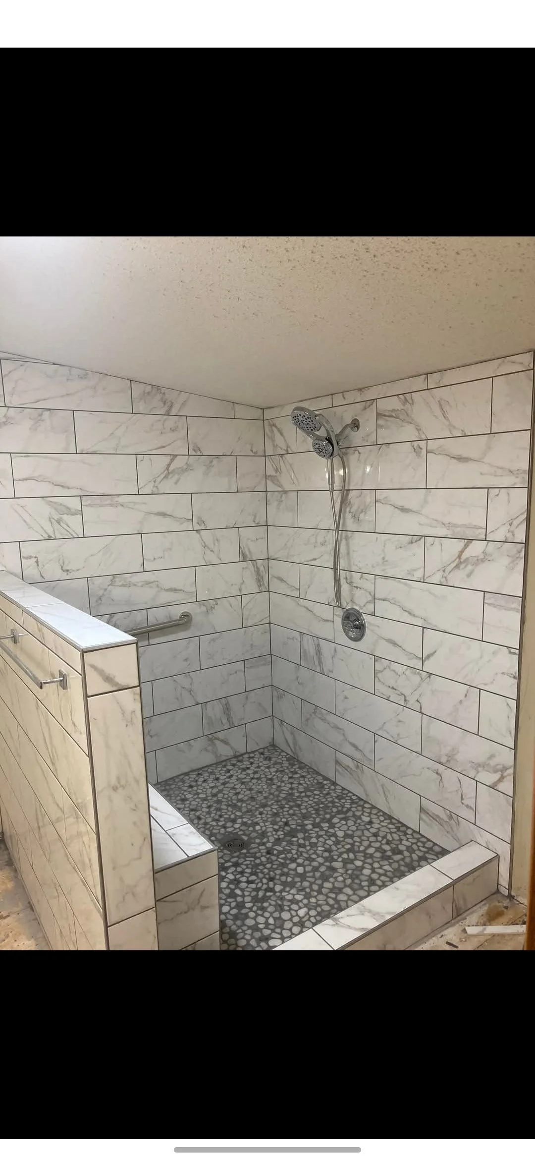 A walk-in shower with marble tile walls, pebble floor, and a handheld showerhead, featuring a partial glass barrier with a towel bar.