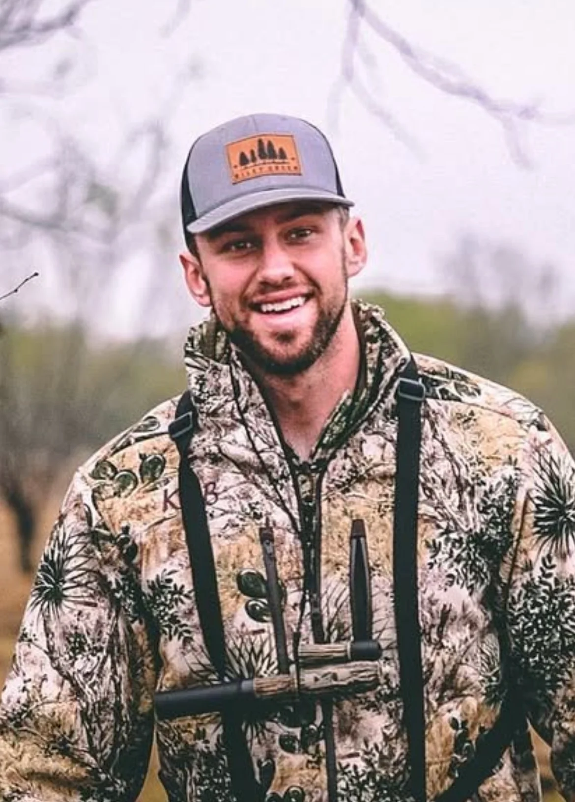 A man smiling outdoors, wearing a camouflage jacket, a gray and black cap, and a shotgun in front of him.