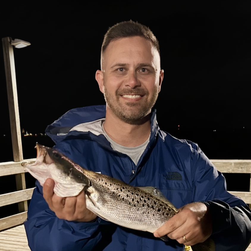Man holding a fish on a wooden pier at night, smiling, wearing a blue windbreaker.