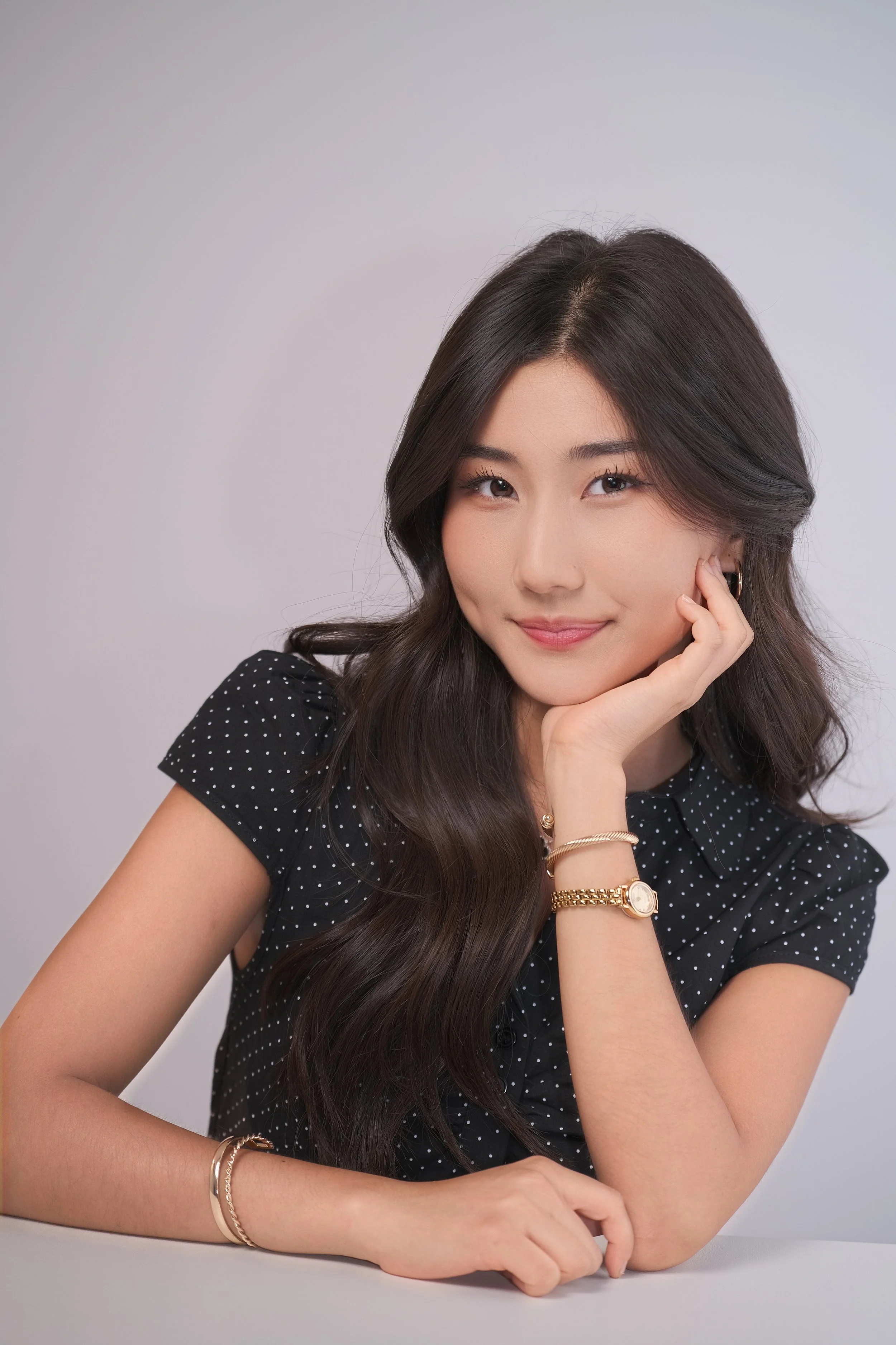 A woman with long dark hair smiling and resting her head on her hand, wearing a black polka dot top and gold jewelry, sitting against a plain background.