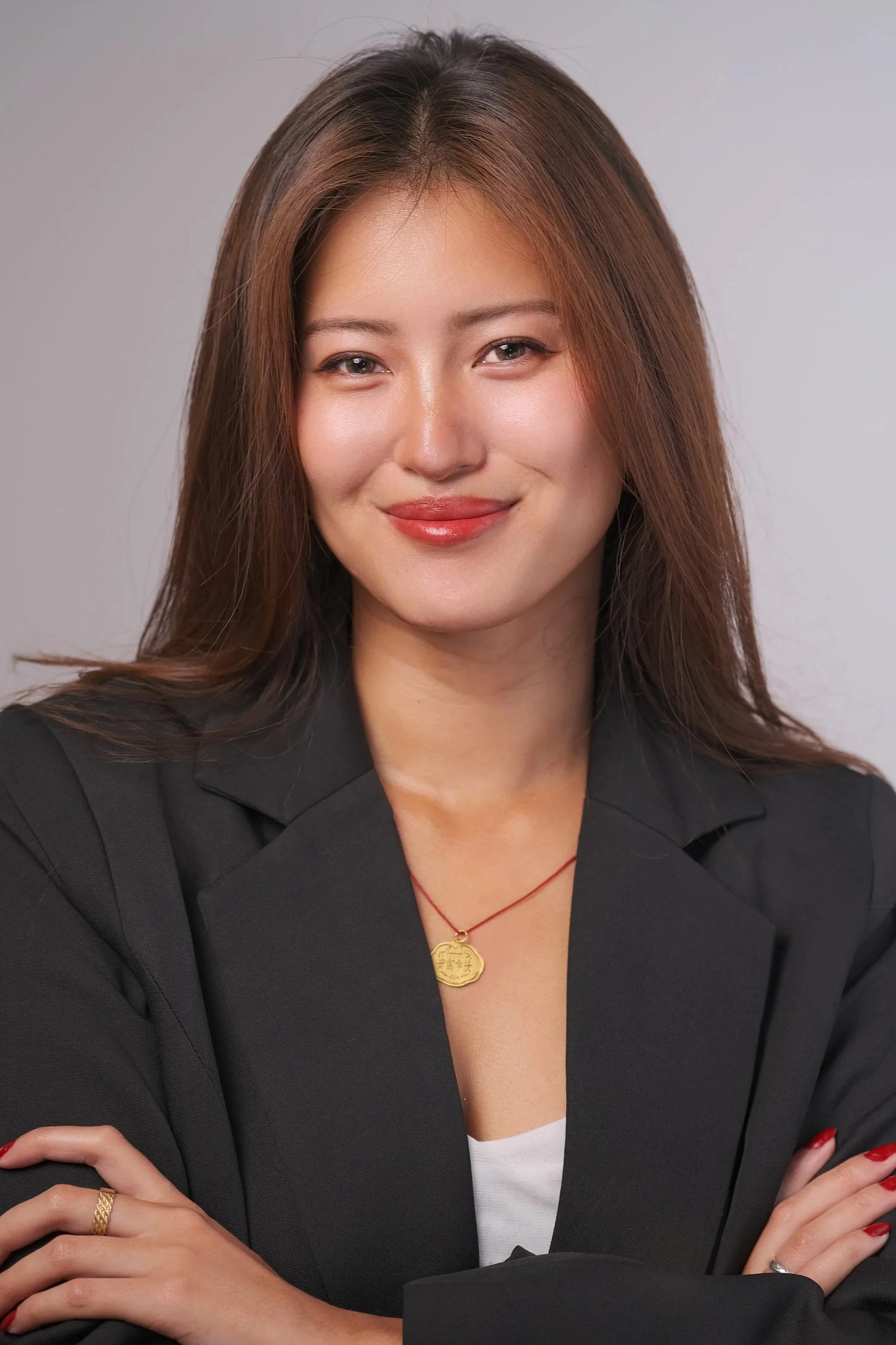 A woman with long brown hair, wearing a black blazer and a white top, smiling with crossed arms, against a neutral background.