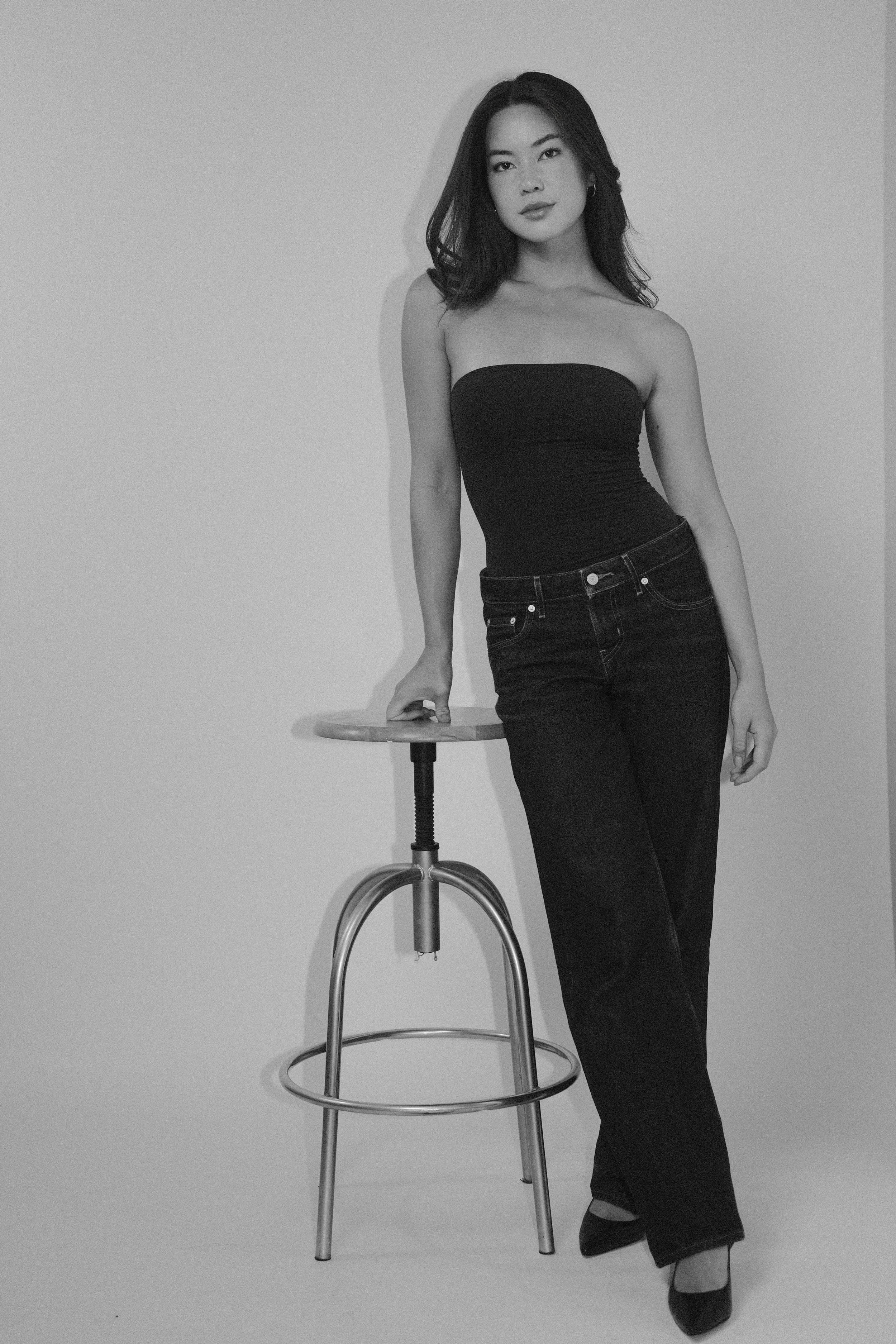 Black and white photo of a young woman with shoulder-length hair, wearing a strapless top and jeans, standing next to a stool with her hand resting on it, against a plain background.