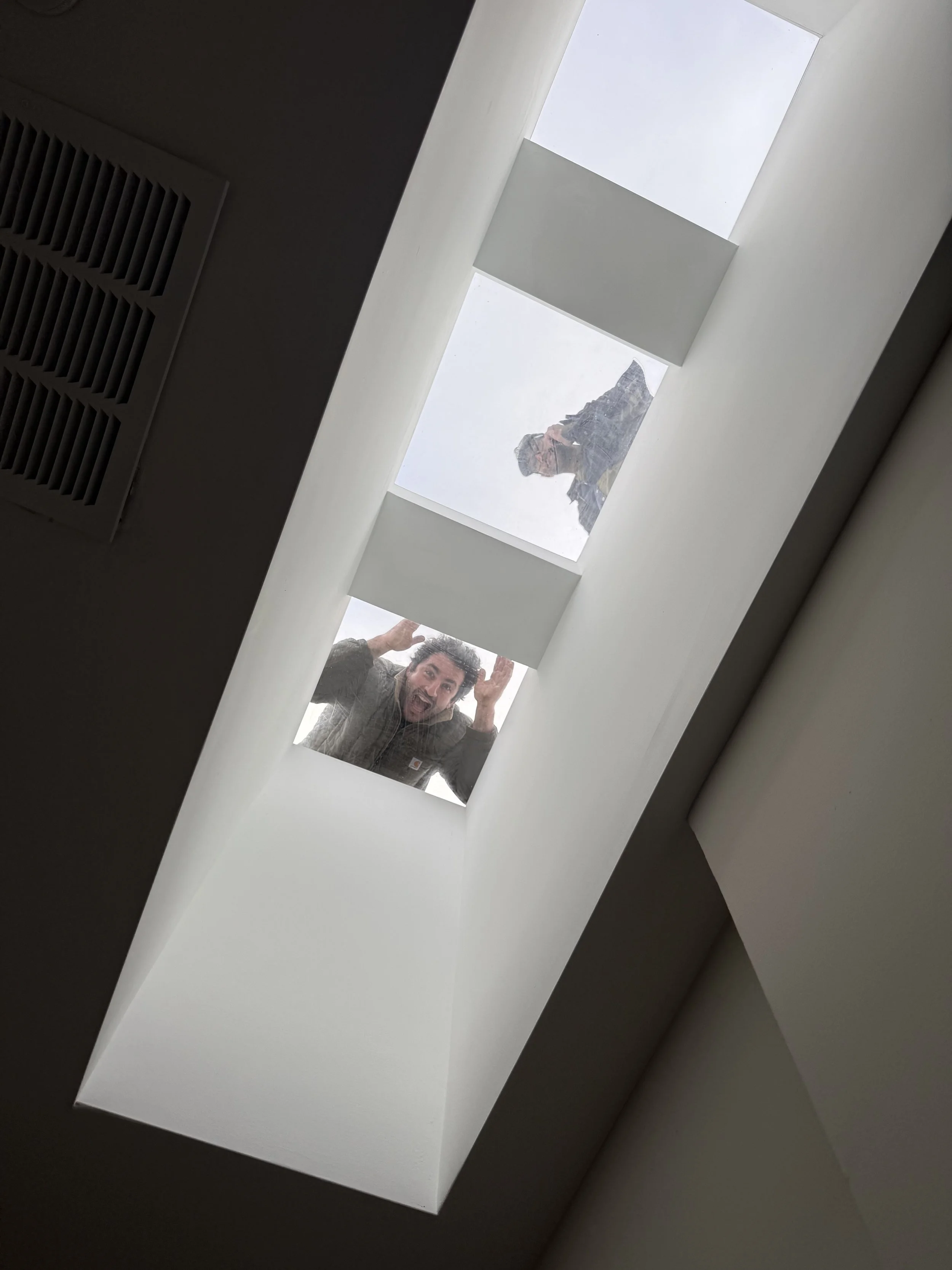 Two people looking down through a skylight window, one of them making a goofy face and the other standing in the background outside the window.