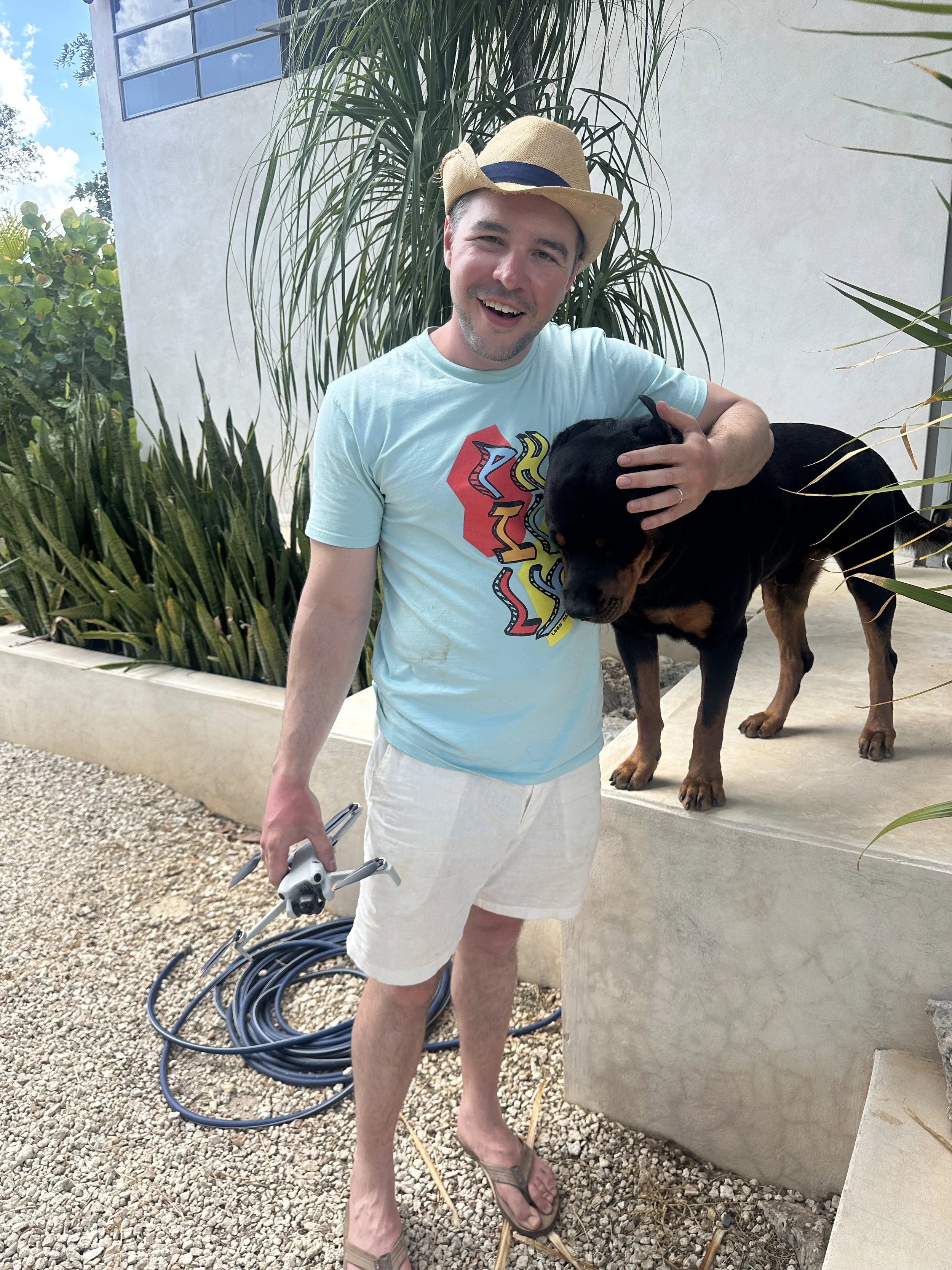 Man wearing a straw hat, light blue graphic t-shirt, white shorts, and flip-flops holding a small black and tan dog, with another larger dog standing on the elevated surface beside him, in an outdoor area with plants and a white building.