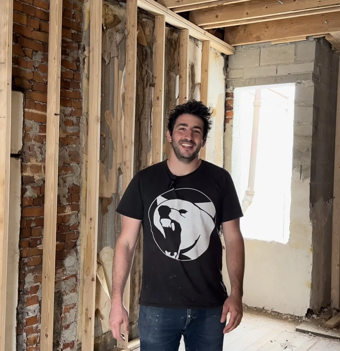 A man with dark, curly hair and a beard smiling indoors during construction or renovation work. He is wearing a black T-shirt with a white graphic of a roaring beast and jeans. The background shows exposed brick and wooden framing in a partially built or renovated space.