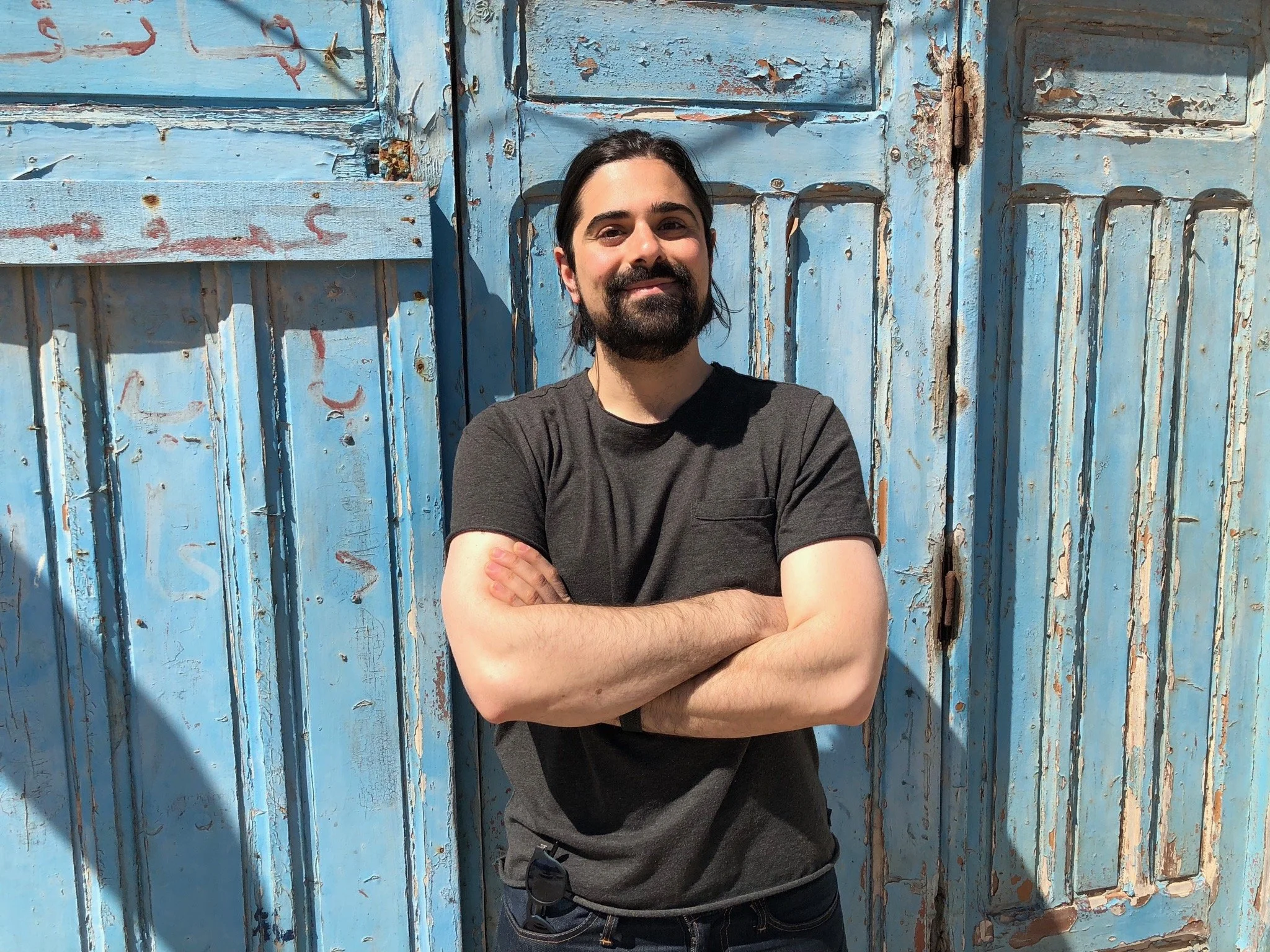 A man with dark hair, a beard, and tattoos on his arms, standing with arms crossed, smiling in front of a weathered blue wooden door.