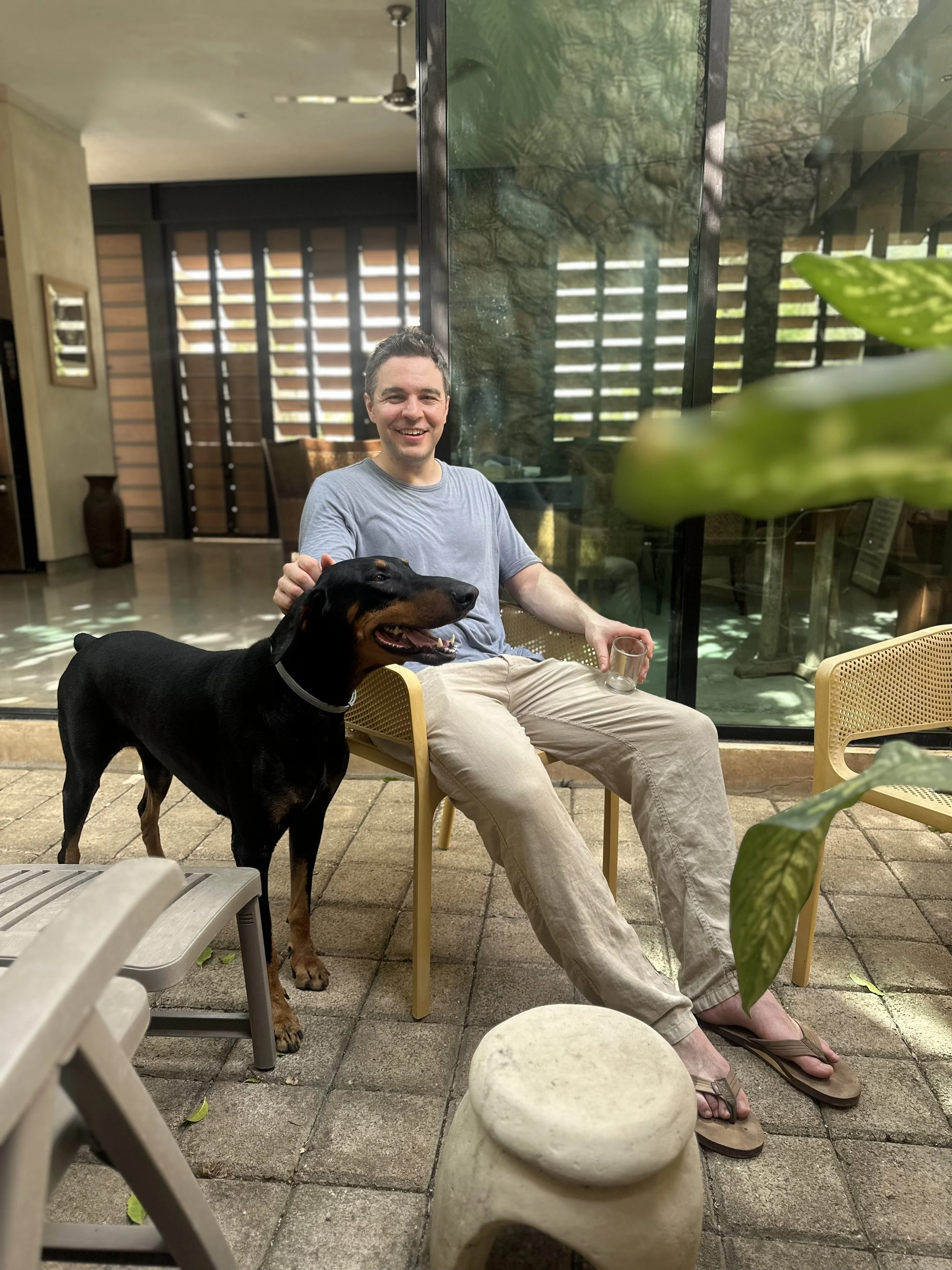 A man sitting outdoors on a yellow chair, smiling, with a black and tan dog standing beside him. The man is wearing a gray t-shirt, beige pants, and flip-flops. The setting appears to be a patio with stone flooring, surrounded by plants and a glass wall, with a wooden fence visible behind.