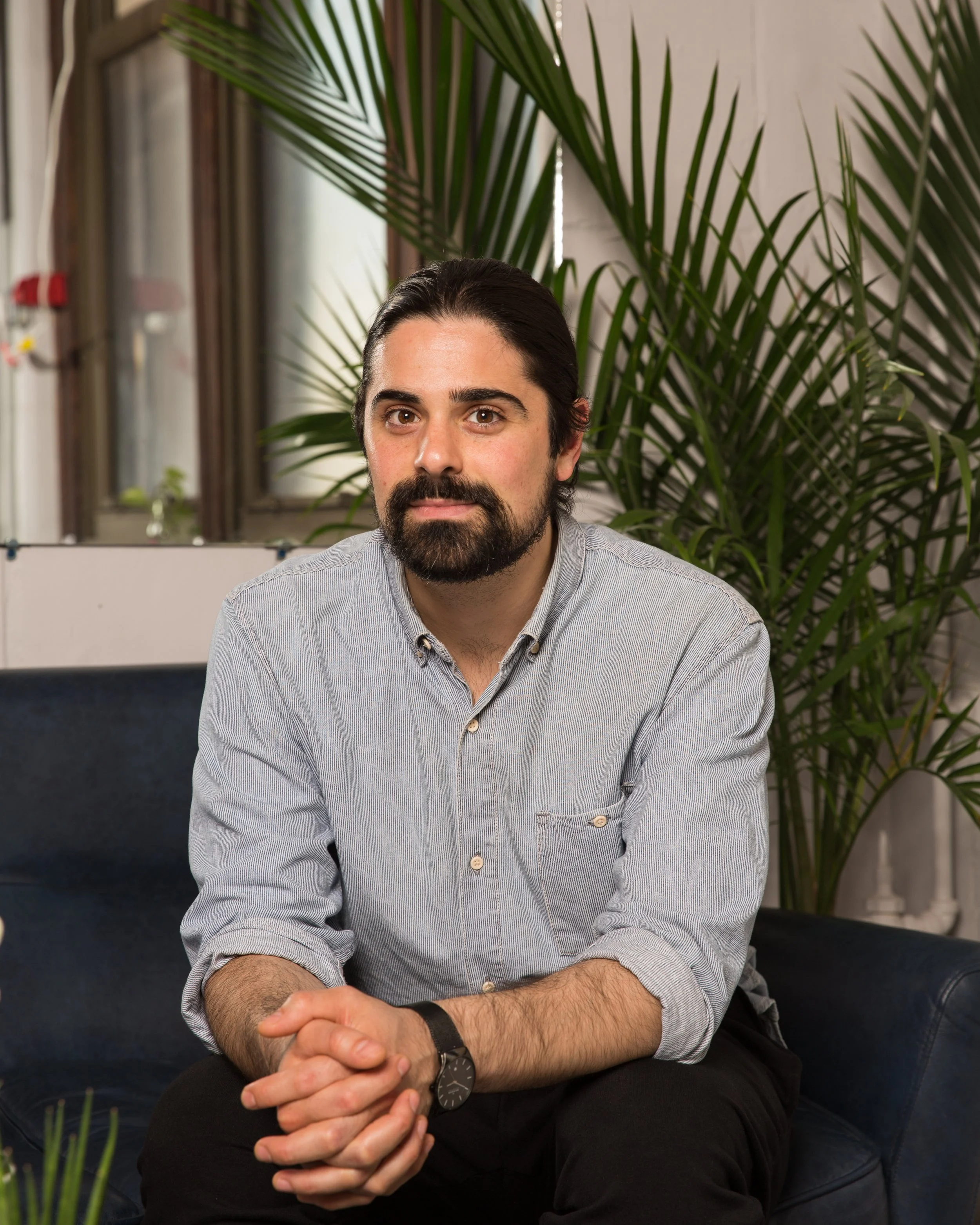 A man with dark hair and a beard sitting on a blue couch, wearing a light gray button-down shirt and a watch, with large green plants and windows in the background.