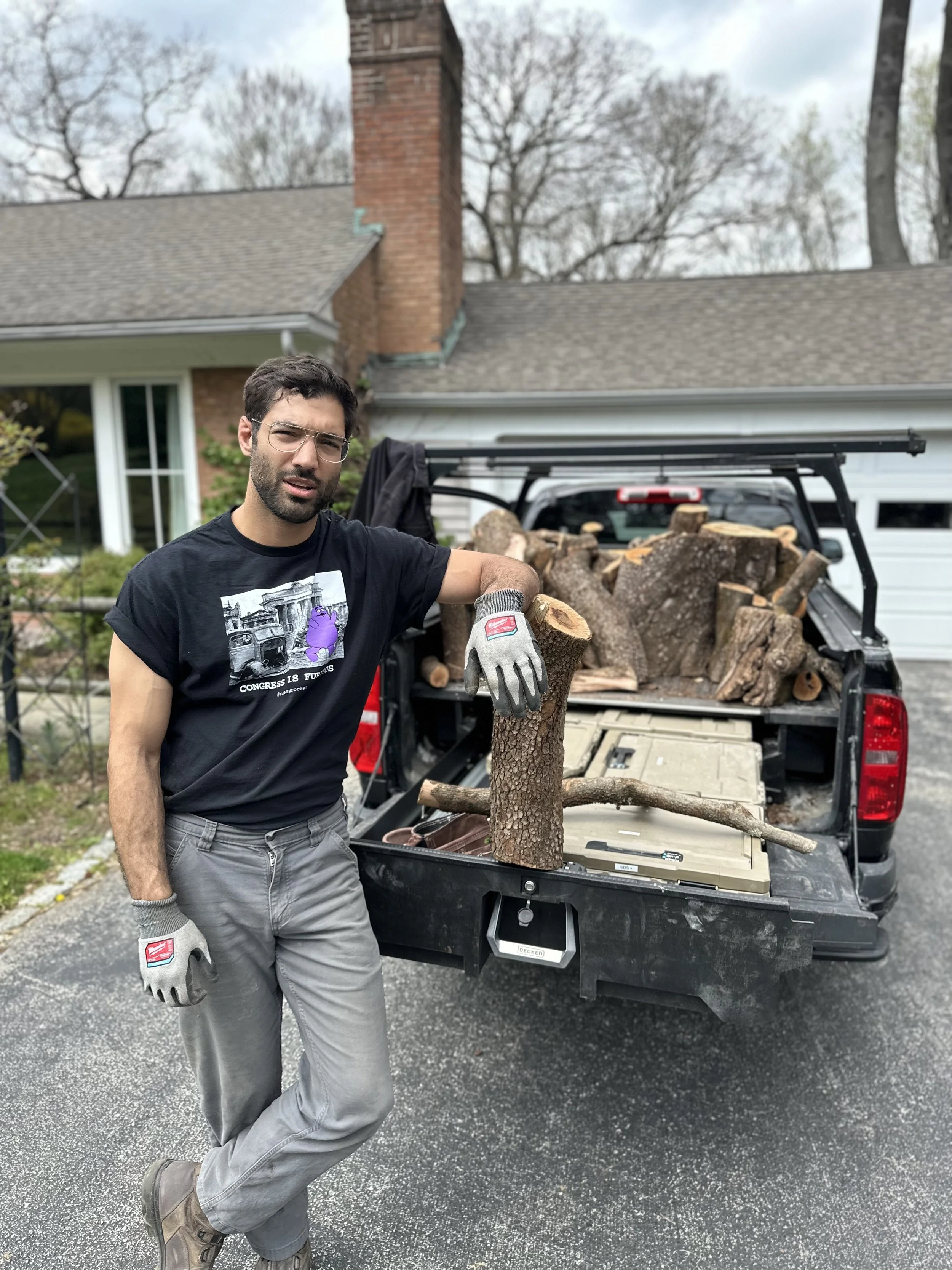 Man in black t-shirt with purple graphic and gray pants standing next to a truck bed full of chopped firewood outside house.