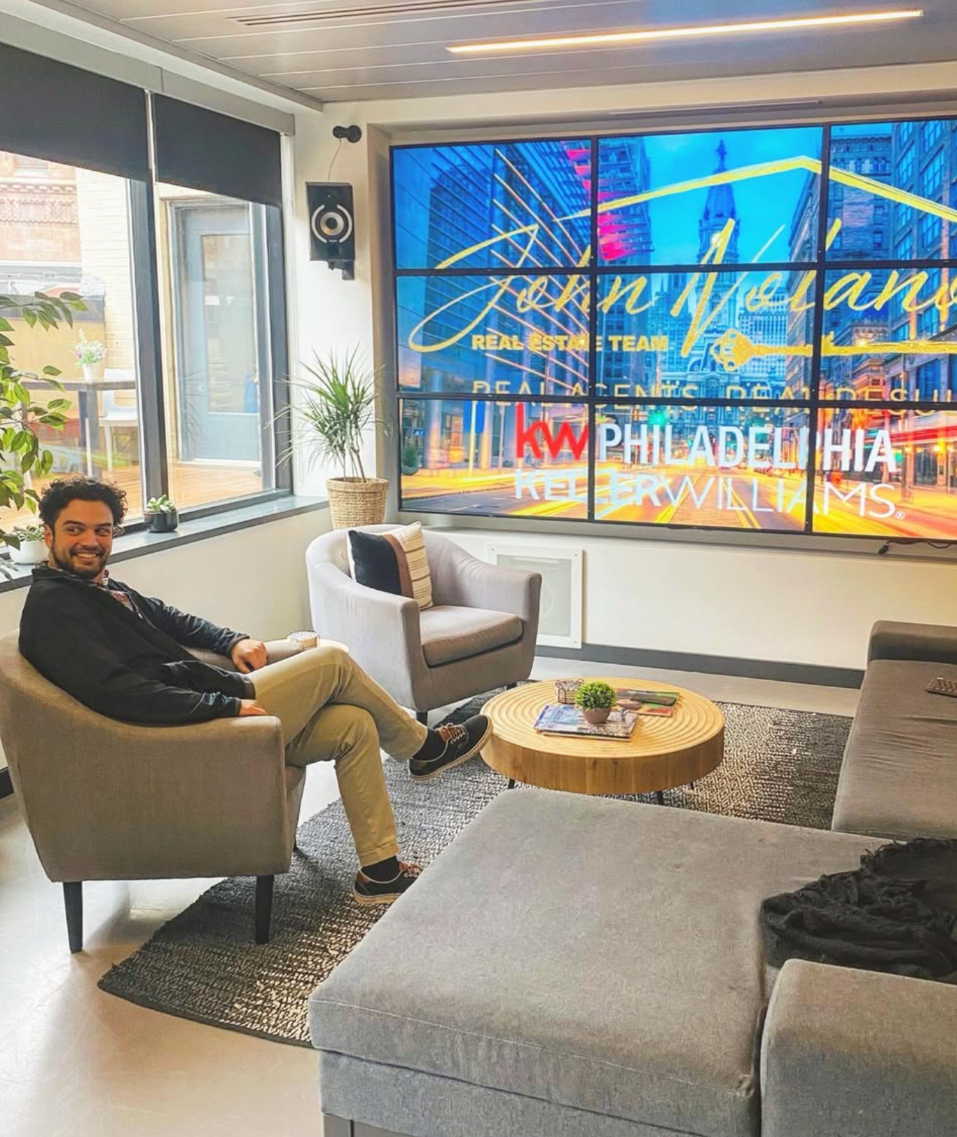 A man sitting on a beige armchair in an office lobby, with a large window behind him and a colorful cityscape on a digital screen displaying real estate company names.