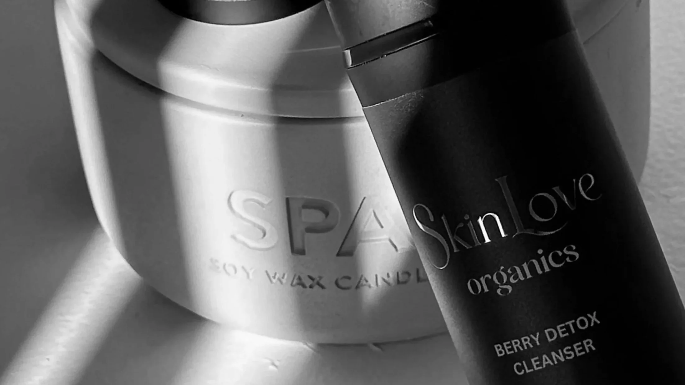 A black bottle labeled 'Skinfove organics Berry Detox Cleanser' leaning against a white candle labeled 'Spa Soy Wax Candles' in a minimalist setting.