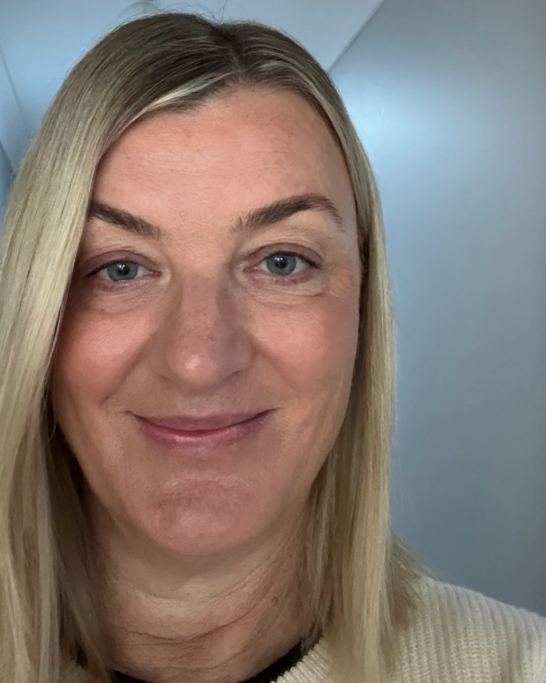 Close-up of a woman with blonde hair, blue eyes, and light skin, smiling softly, wearing a cream-colored sweater.
