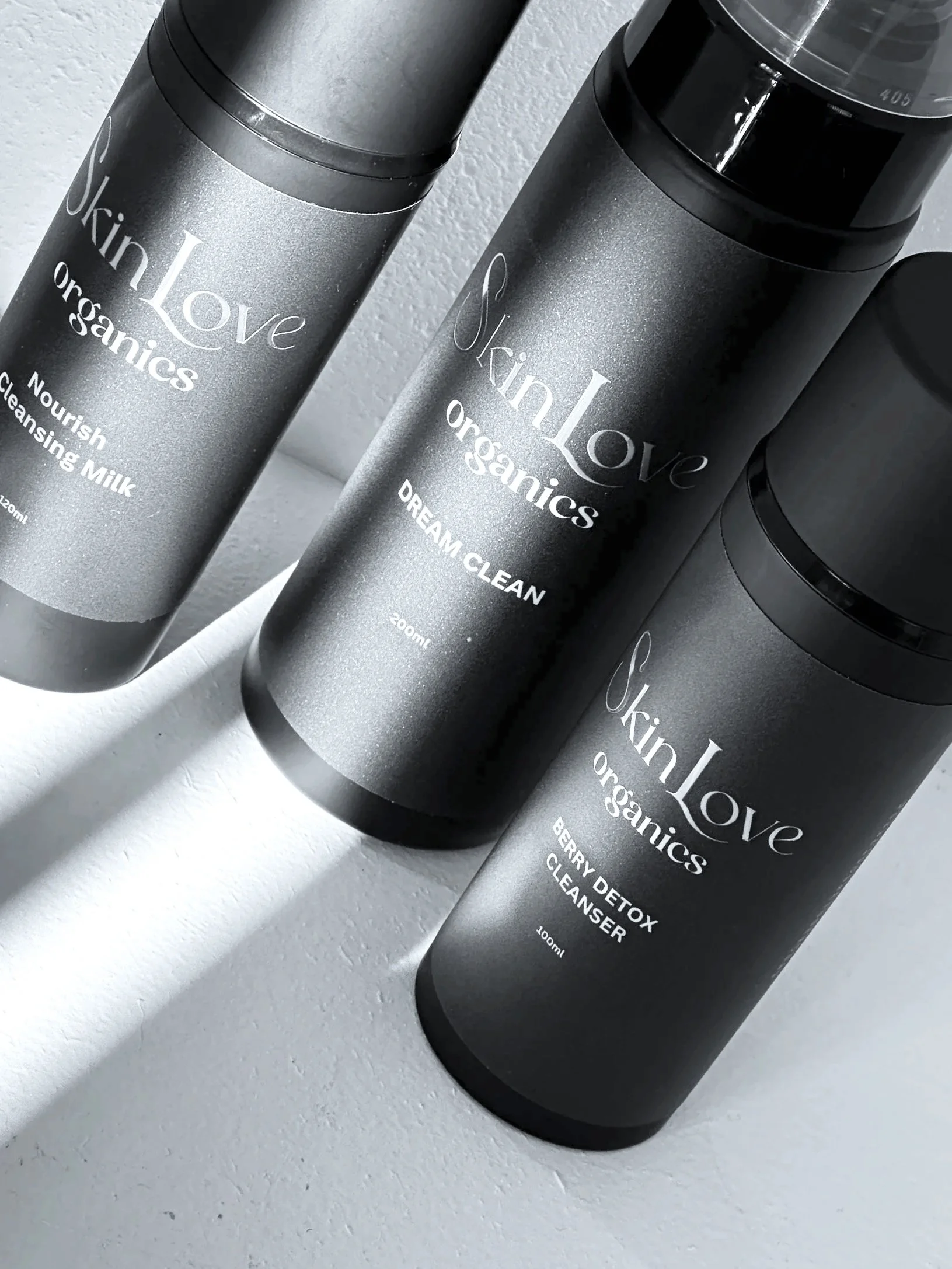 Three black bottles of SkinLove Organics skincare products on a white surface. The middle bottle is labeled 'Dream Clean,' the right one 'Berry Detox Cleanser,' and the left one 'Nourish Cleansing Milk.'