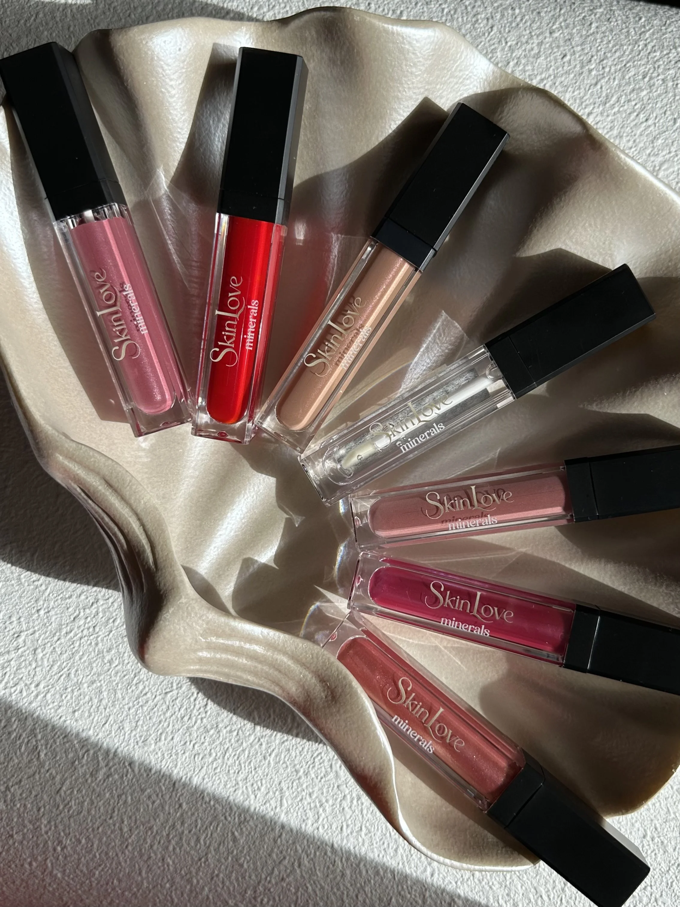 Collection of six SkinLove Minerals lip glosses in various shades, arranged in a fan shape on a wavy, cream-colored dish.