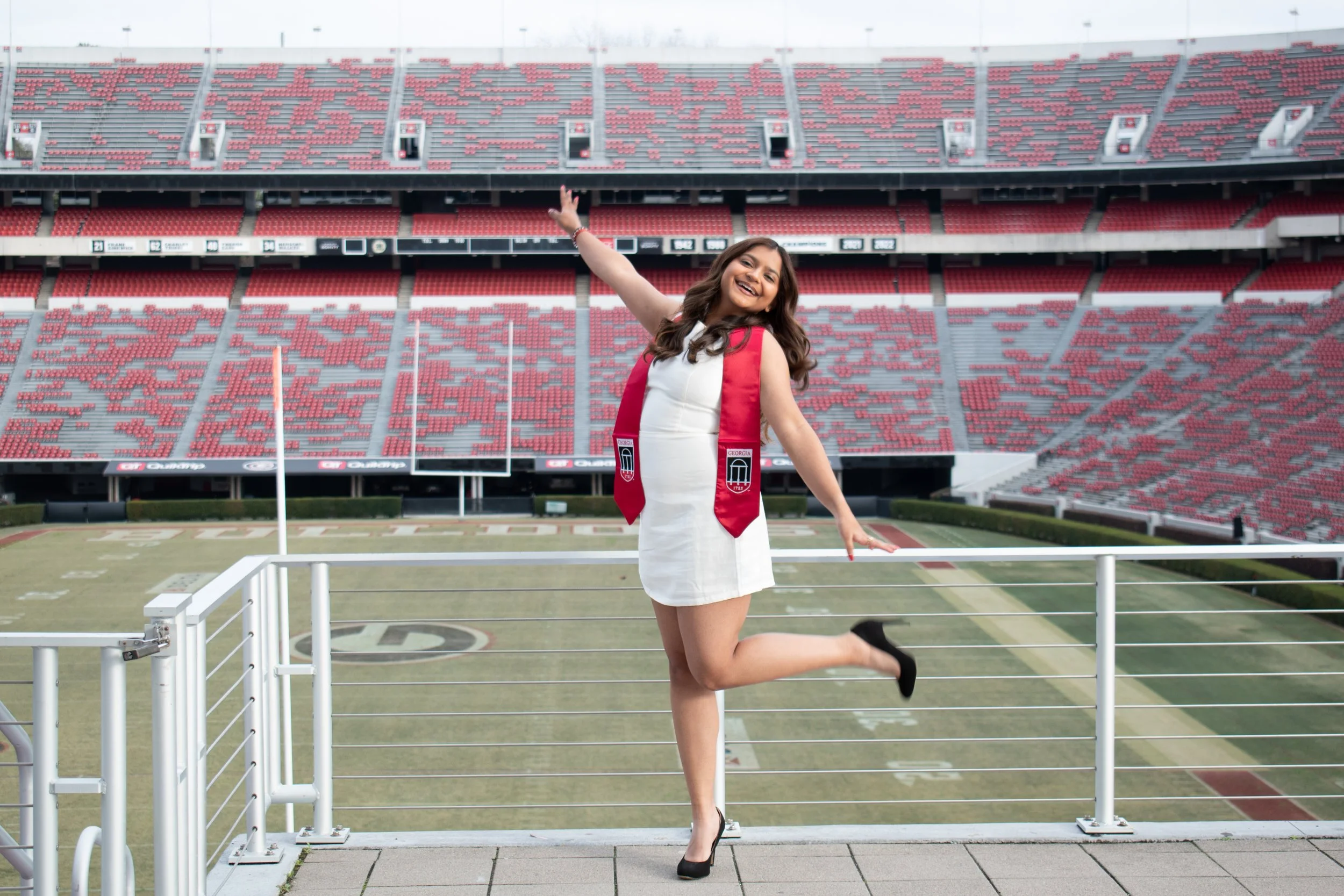 Excited 2026 Senior student/graduate at The University of Georgia Sanford Stadium, wearing a white dress, black heels, and a red Dawgs sash, standing on a platform jumping and smiling at the camera