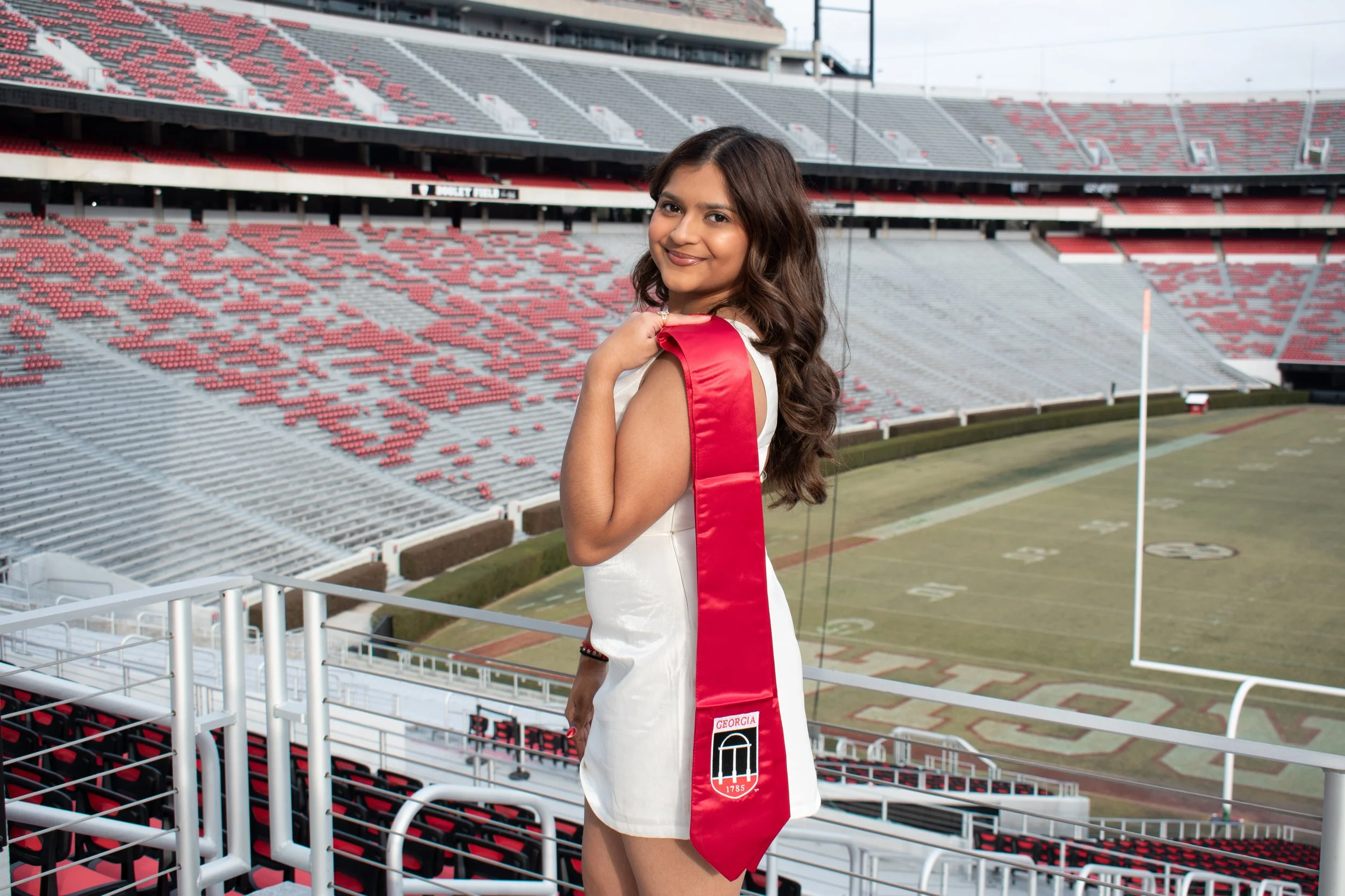 A woman in a white dress with a red sash draped over her shoulder, standing in a stadium with empty red and gray seats, smiling and looking at the camera.