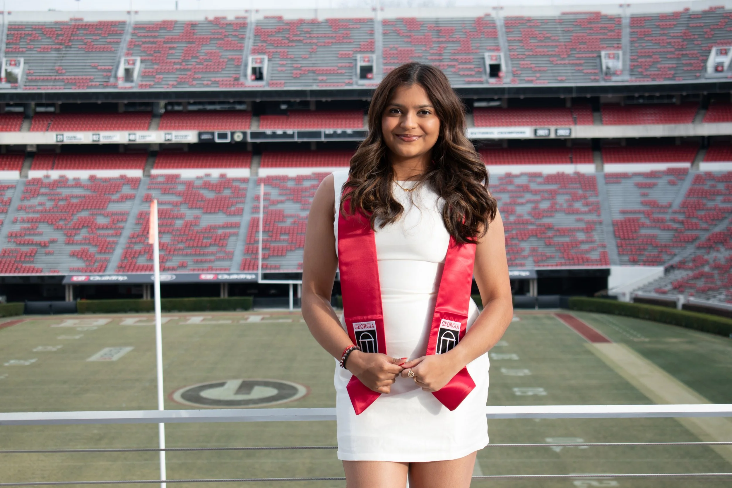 Excited 2026 Senior student/graduate at The University of Georgia in front of Sanford Stadium in a white dress and red Dawgs sash