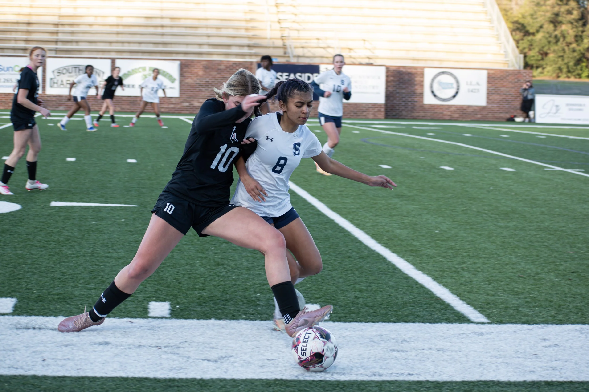 Two female soccer players competing for ball on field, with more players in background, both running trying to get control of ball