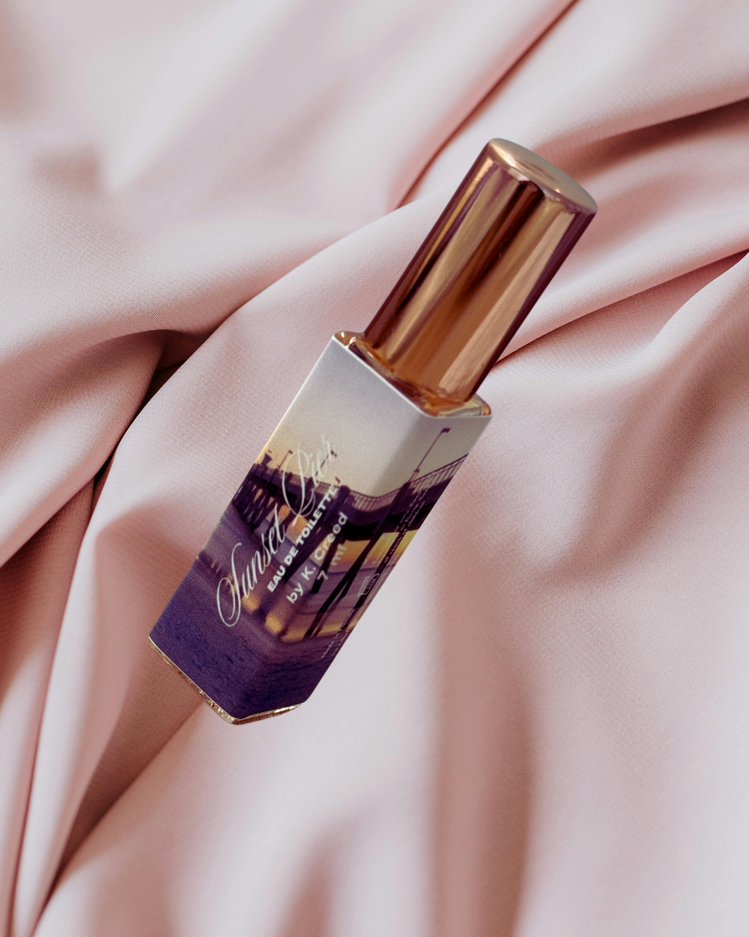 A perfume spray bottle with a golden cap, resting on pink satin fabric. The label shows an image of a bridge and a sunset, and the text reads "Ondine Eau de Toilette by K. Flor".