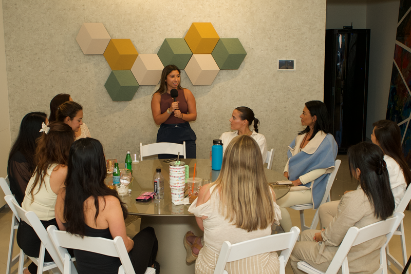 A woman giving a talk to a group of women seated around a table in a conference room with hexagonal wall decorations.