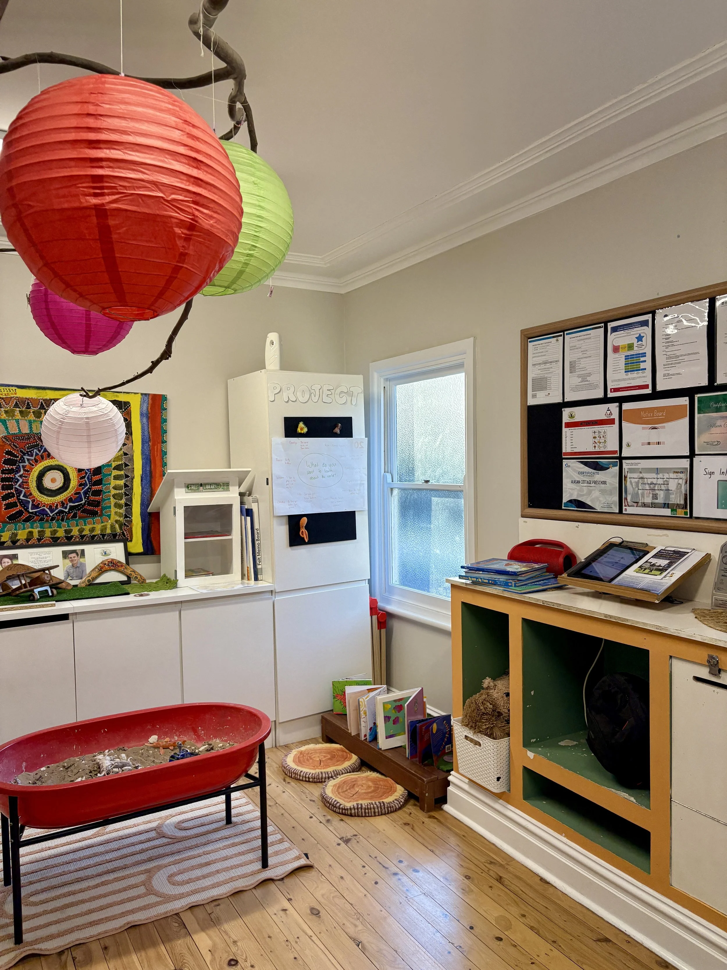 Colorful paper lanterns hanging from a branch in a classroom with educational materials, books, and a sandbox.