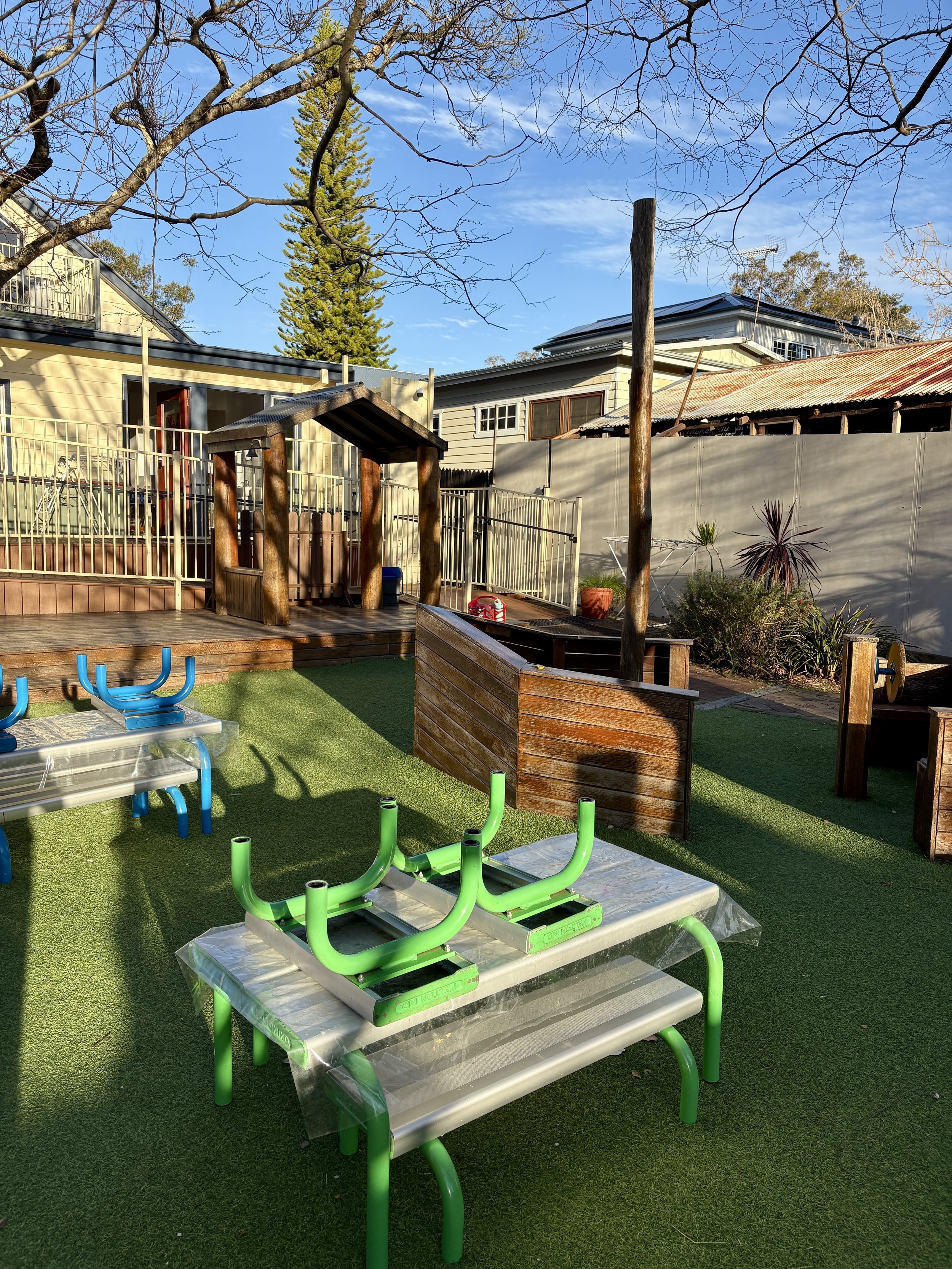Outdoor playground area with green grass flooring, relocated tables, a playhouse, and surrounding trees and buildings.