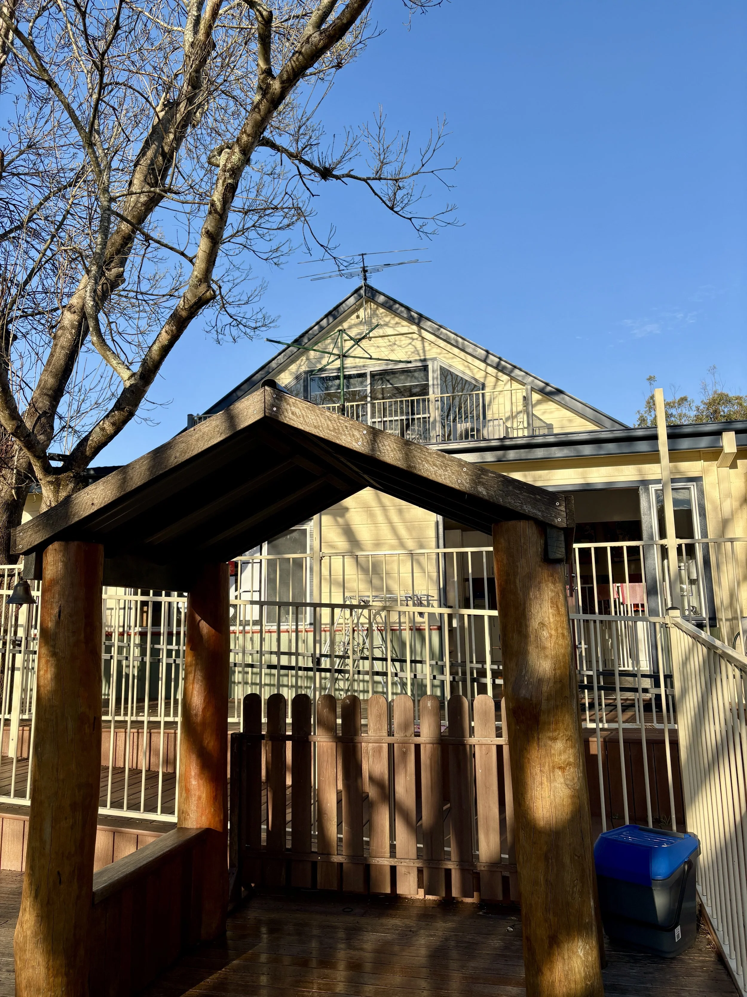 A residential house with a triangular roof, yellow exterior, and multiple decks with railings. There is a leafless tree in the foreground, a blue sky in the background, and a wooden exit gate in the lower part of the image.