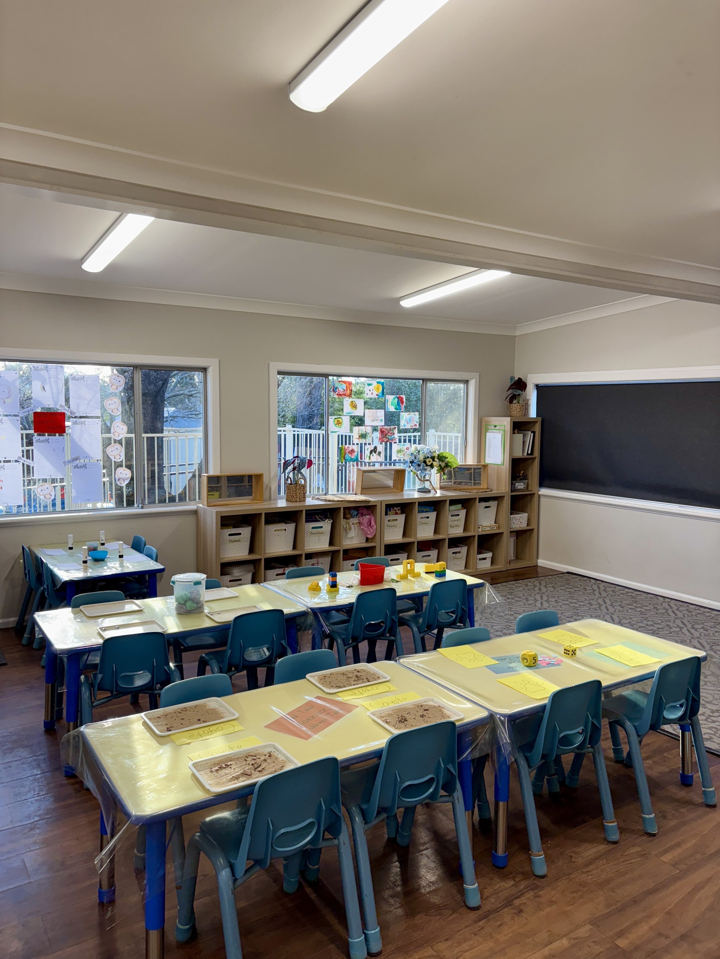 A brightly lit classroom with three yellow tables, each covered with plastic tablecloths and food trays. Small blue chairs are arranged around the tables. The classroom has large windows decorated with children's artwork and a blackboard on the wall.