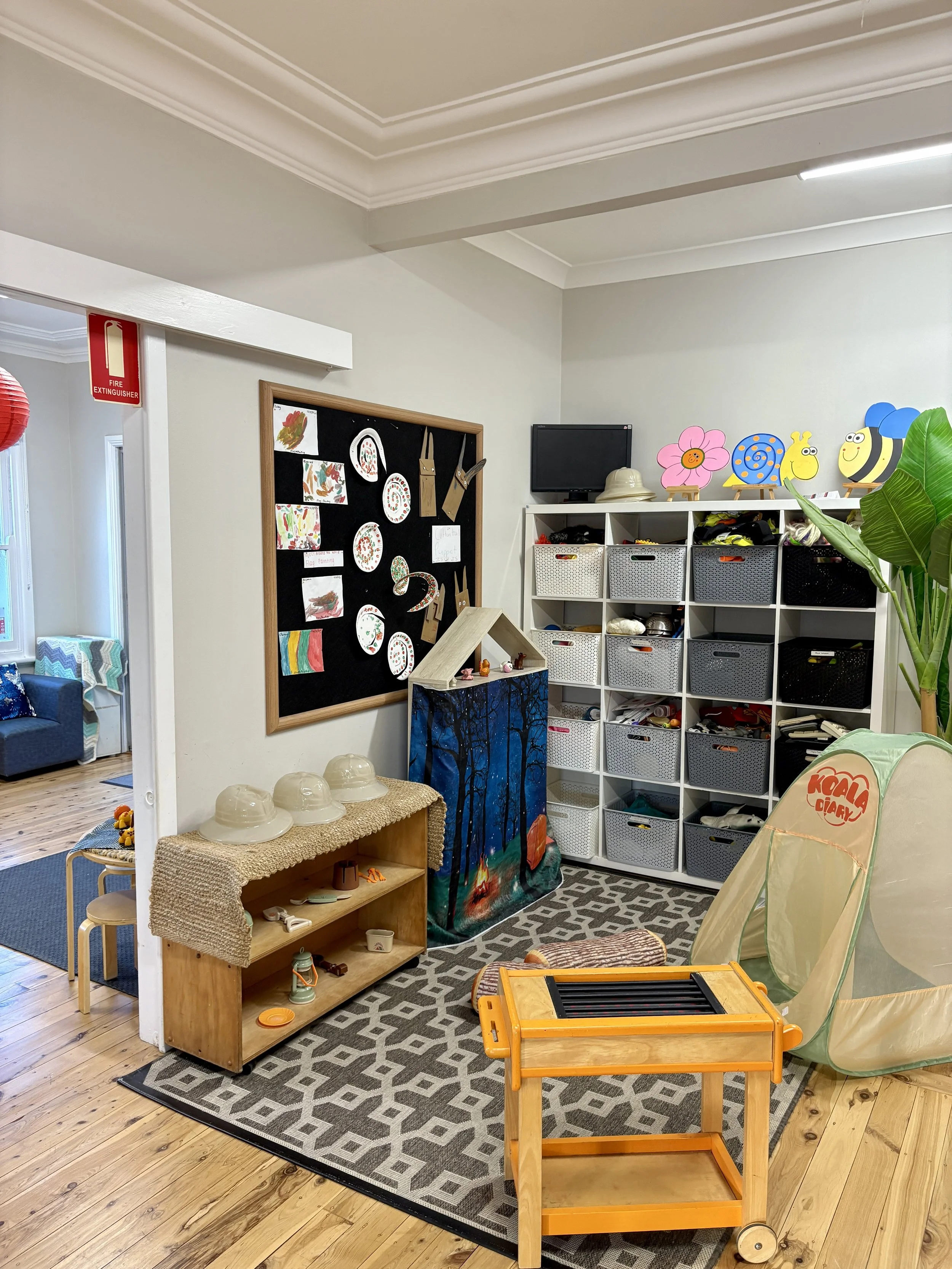 A children's playroom with a blackboard wall, storage cubbies filled with toys, a tent, and a small wooden toy cart.