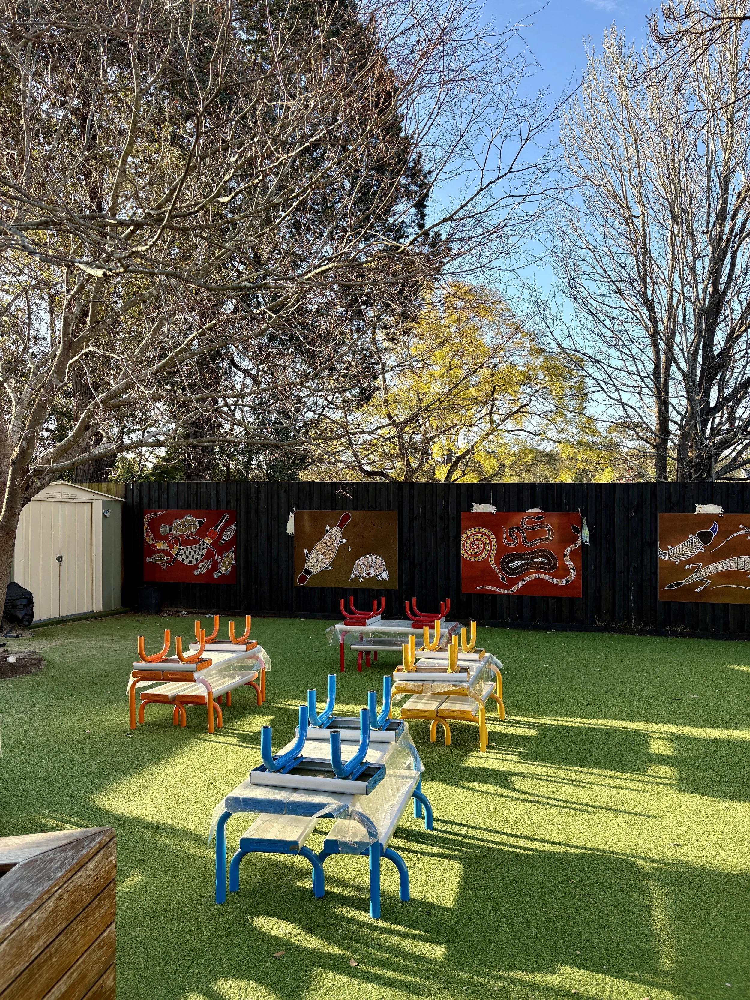 Empty outdoor playground with upside-down benches in blue, yellow, orange, and red, on artificial grass. A black wooden fence with Aboriginal paintings of animals and symbols in red, brown, and white is in the background. Bare trees and a clear blue 