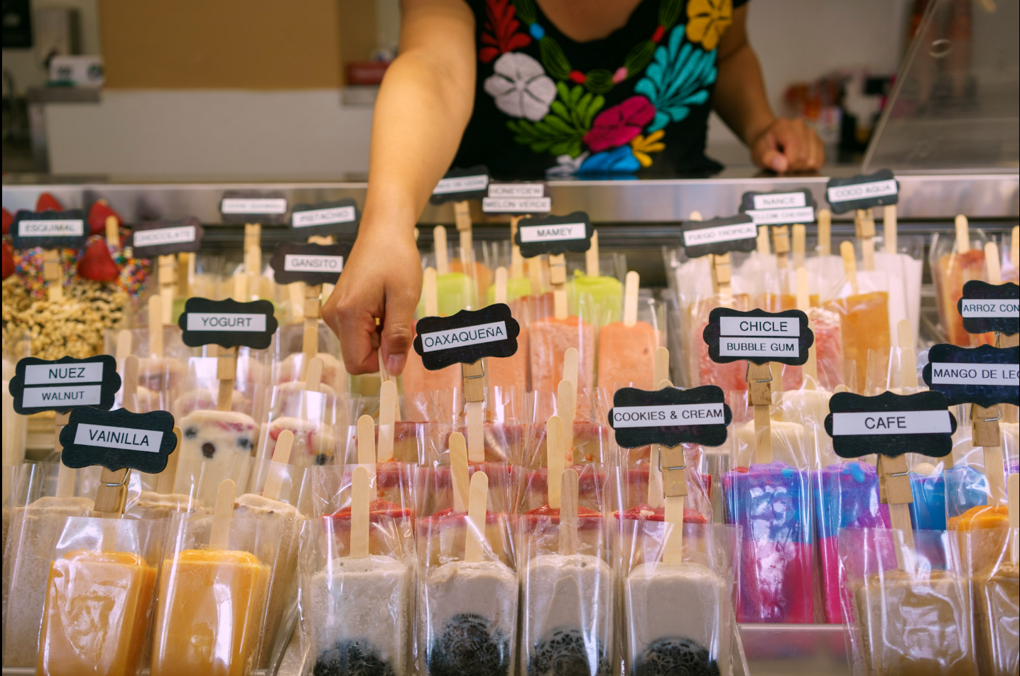 A person selecting colorful ice cream popsicles at a vendor's stall, with labels like vanilla, yogurt, cookies and cream, and bubble gum.