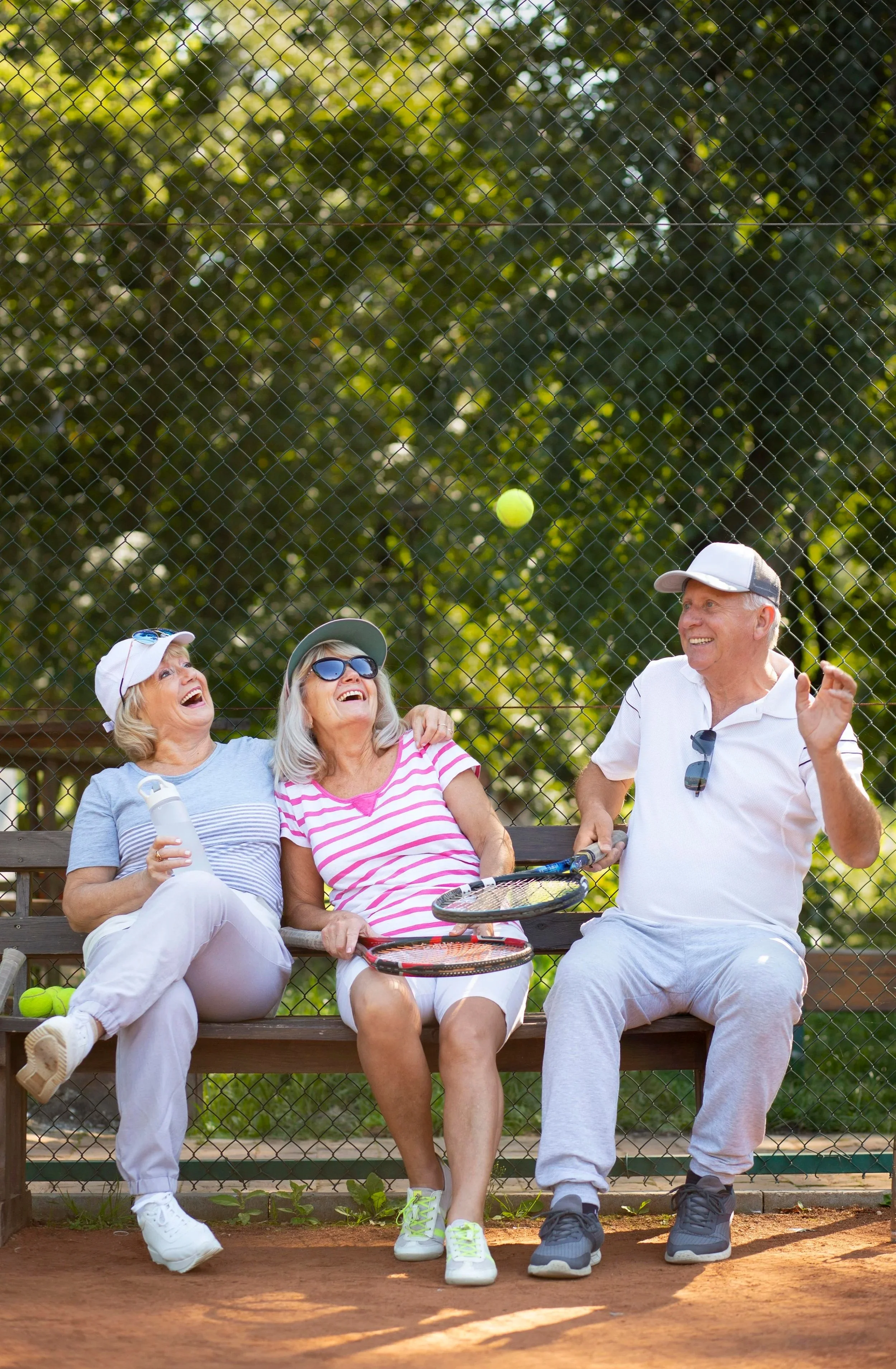 Four older adults sitting on a bench on a tennis court, laughing and chatting, with tennis rackets and tennis balls nearby.