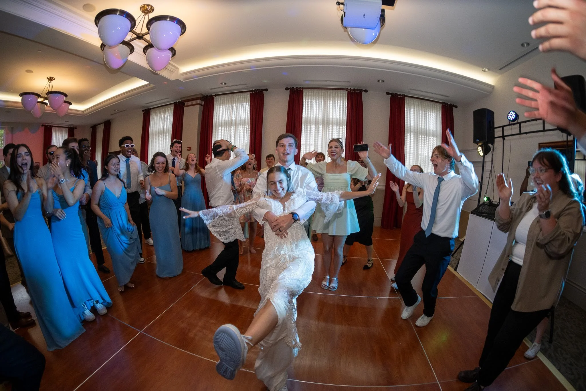 People dancing and celebrating at a wedding reception with the bride and groom in the center, surrounded by guests in colorful dresses and formal wear in a decorated ballroom.
