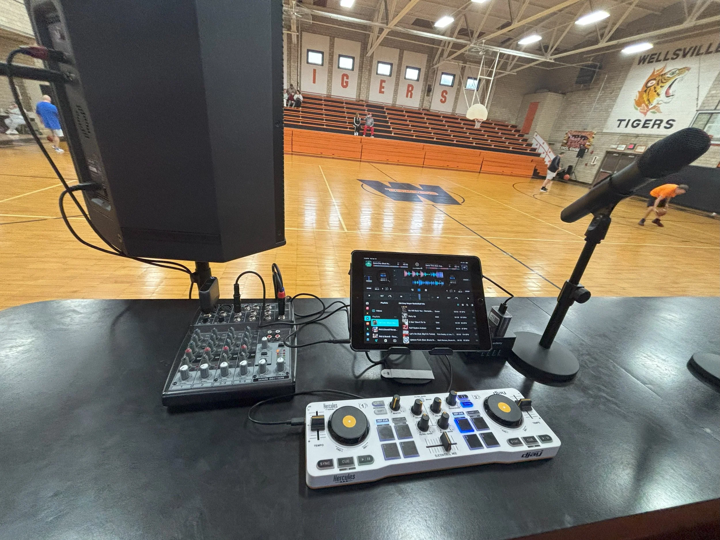 DJ setup on a table in a gymnasium with a speaker, tablet, microphone, mixer, and DJ controller. In the background, people are playing basketball on a court with gym bleachers and a wall decorated with a tiger mascot.