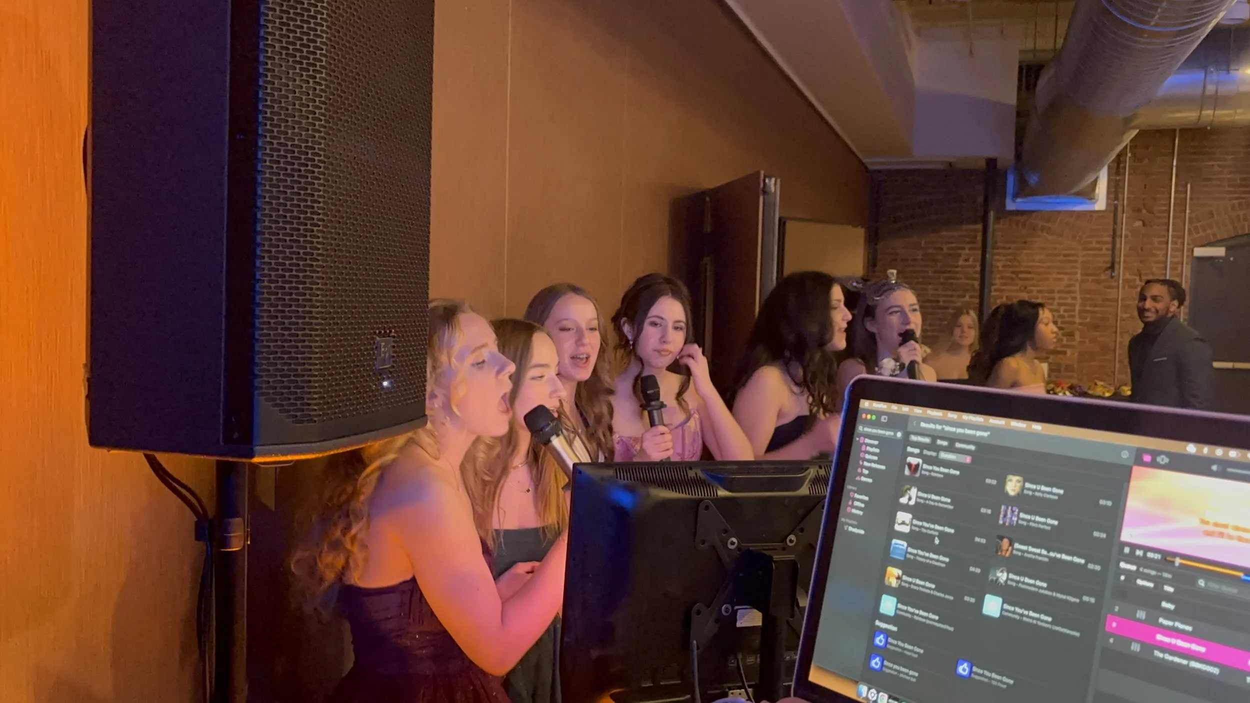 Group of women singing karaoke in a dimly lit room with a brick wall background, a man smiling in the background, and a DJ setup with a computer in the foreground.