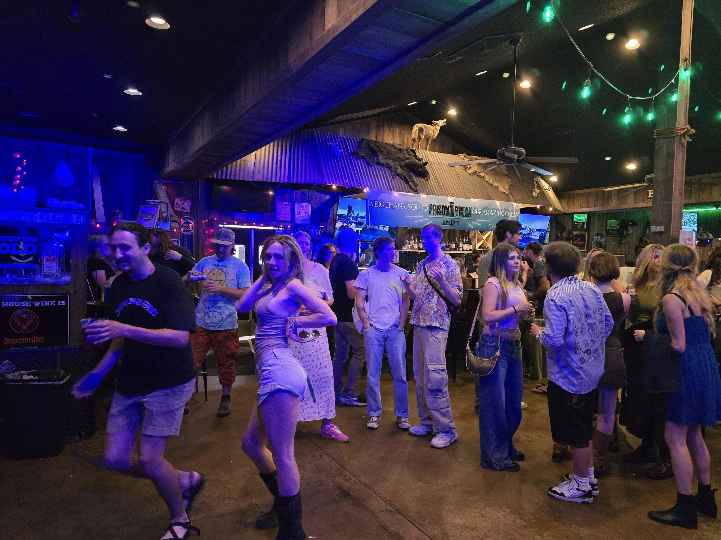 People dancing and socializing inside a bar or club with wooden walls, neon lights, and a mounted animal head and fur decorations on the wall.