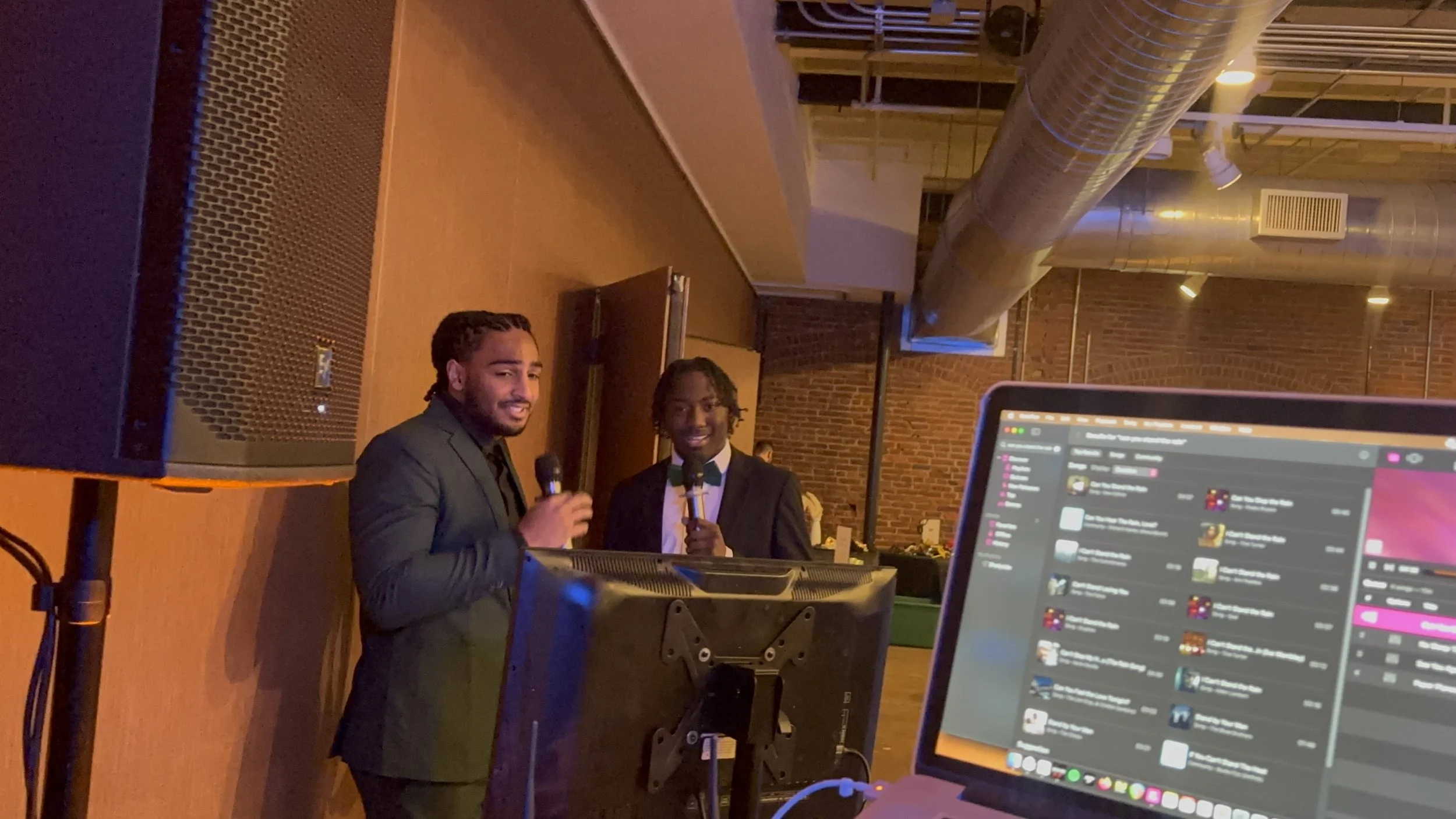 Two men in formal suits holding microphones and smiling, standing behind a DJ booth in a dimly lit venue with brick walls and industrial ceiling ducts, with a computer monitor visible in the foreground.