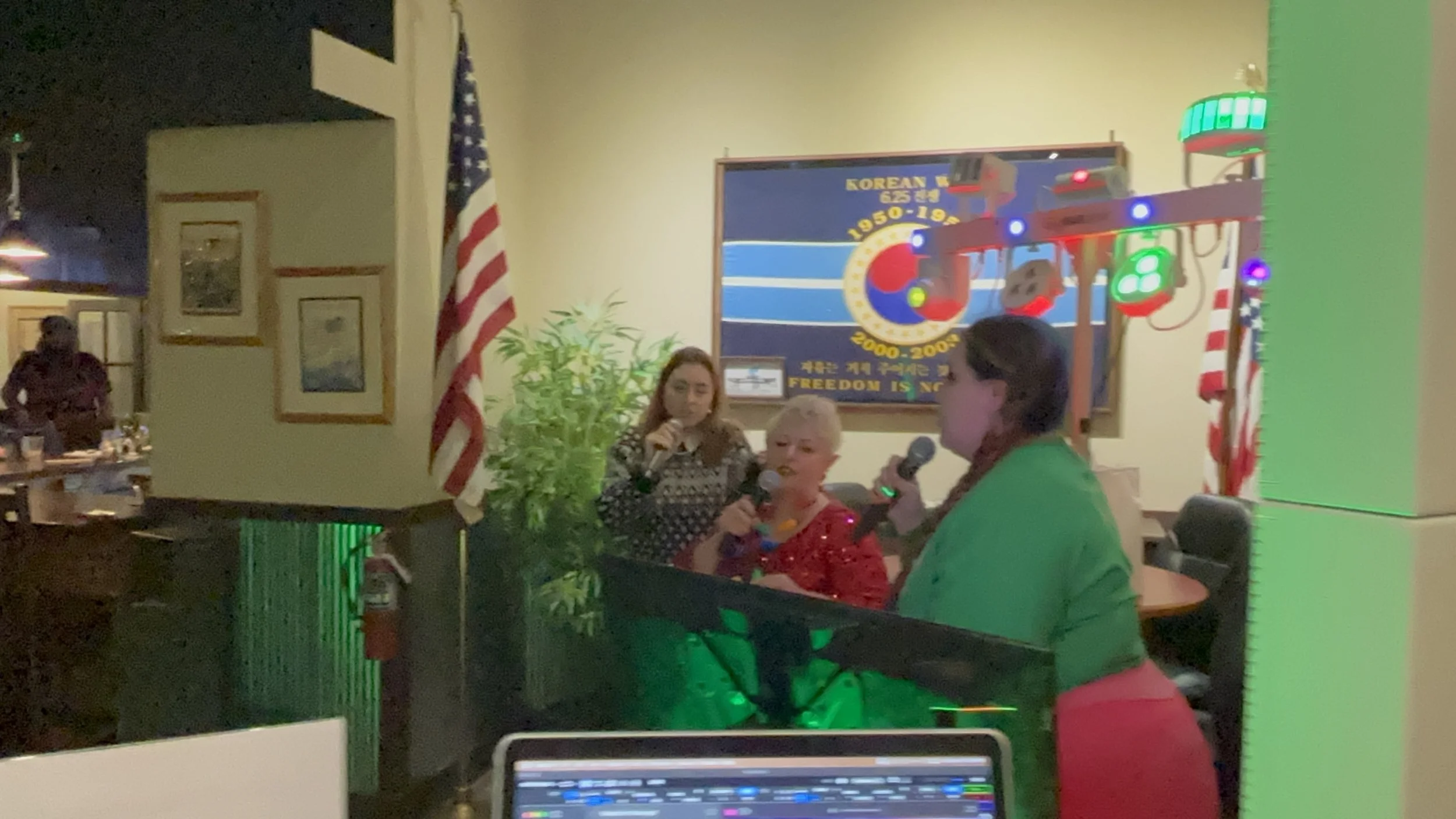 Three women with microphones performing karaoke in a room decorated with American flags and a motivational poster, with one woman in a red blouse and two others in patterned tops.