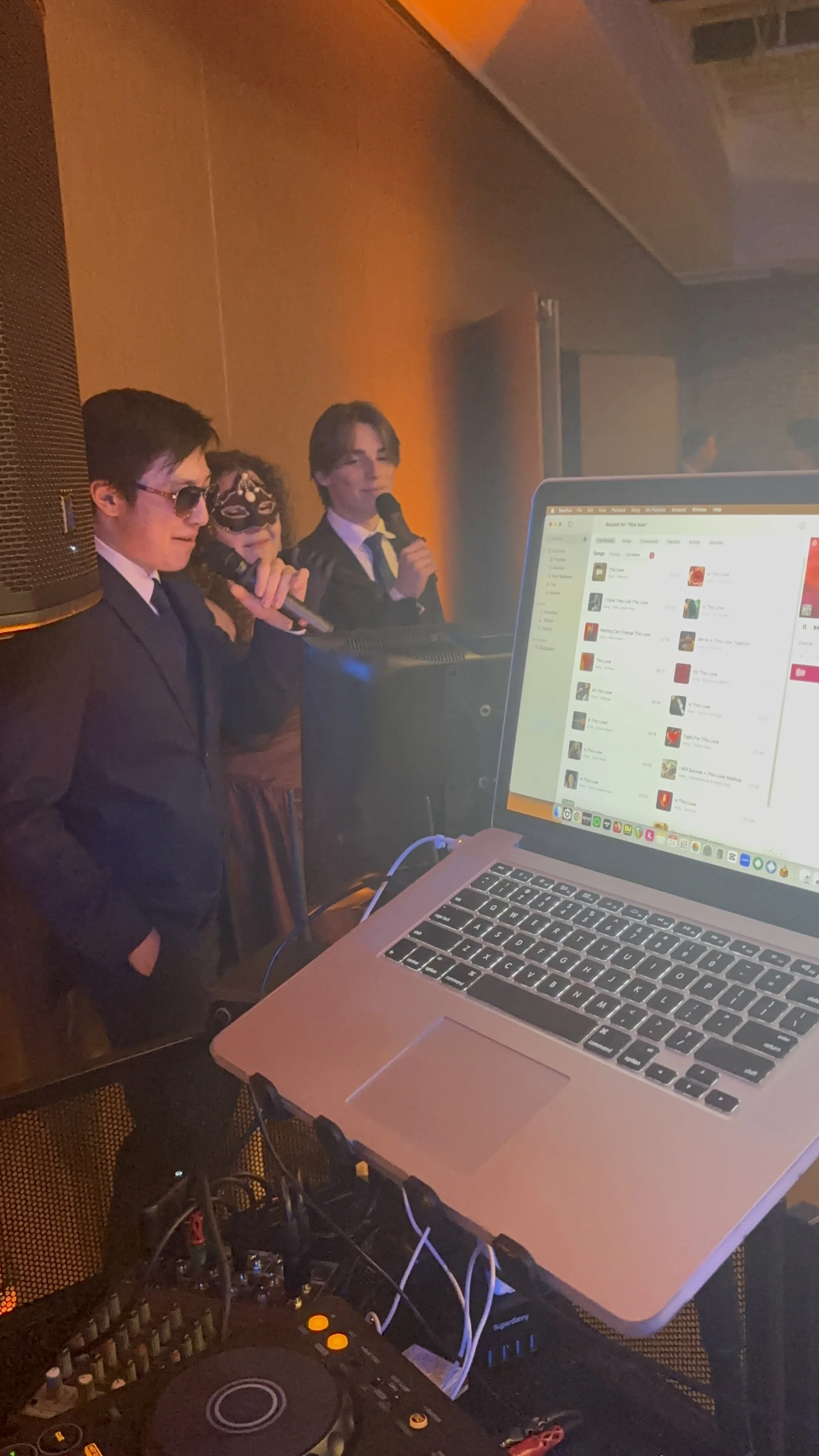 A DJ setup with a laptop, mixer, and headphone, and three people in elegant black suits and dresses, standing on a stage with microphones, likely performing or speaking at an event.