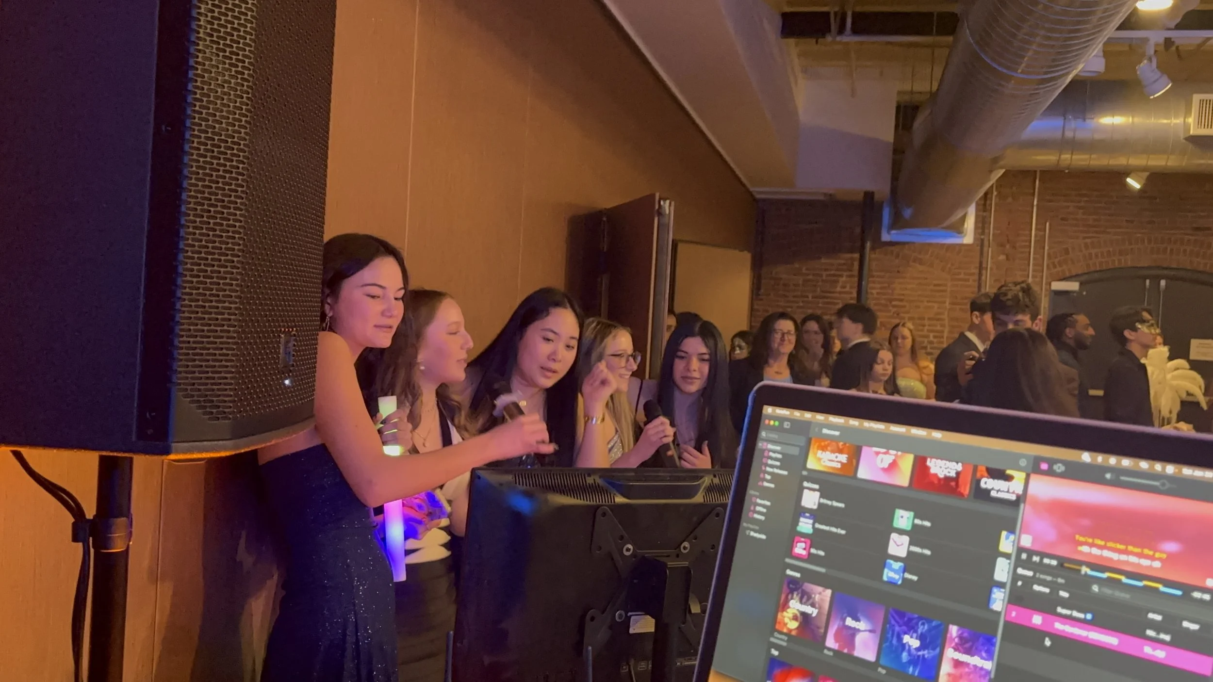 Group of women singing into microphones at an indoor event, with a DJ or music setup in the foreground and a crowd of people in the background.