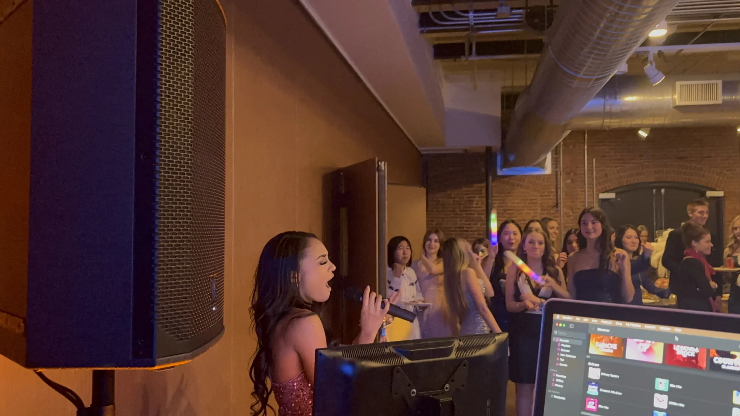 A woman sings into a microphone at a party, surrounded by a group of women holding glow sticks and smiling in a room with brick walls and industrial ductwork.