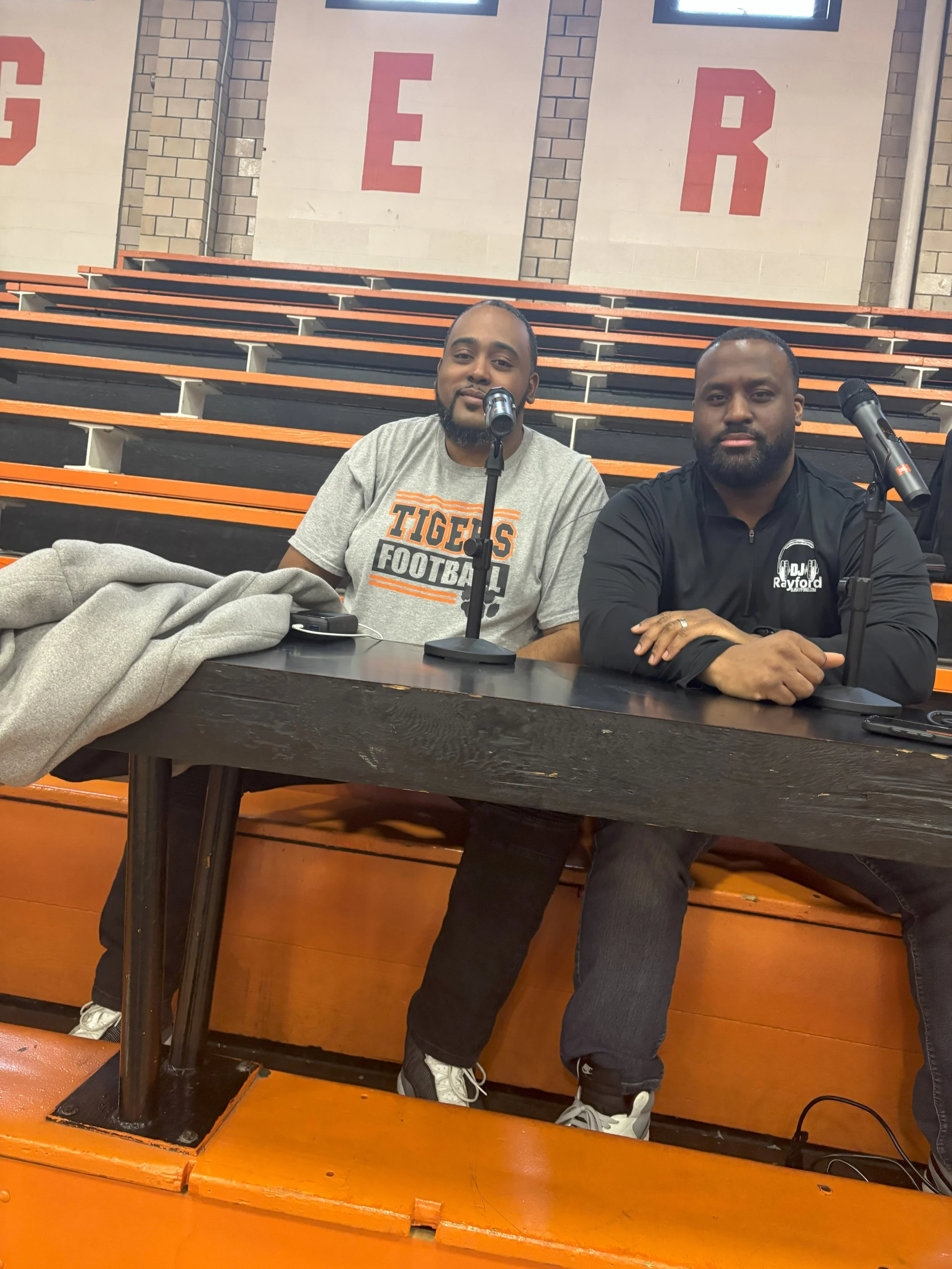 Two men sitting at a table in a sports arena with orange and black bleachers behind them. One is wearing a gray T-shirt with 'TIGERS FOOTBALL' on it, and the other is in a black jacket with a logo. They have microphones in front of them.