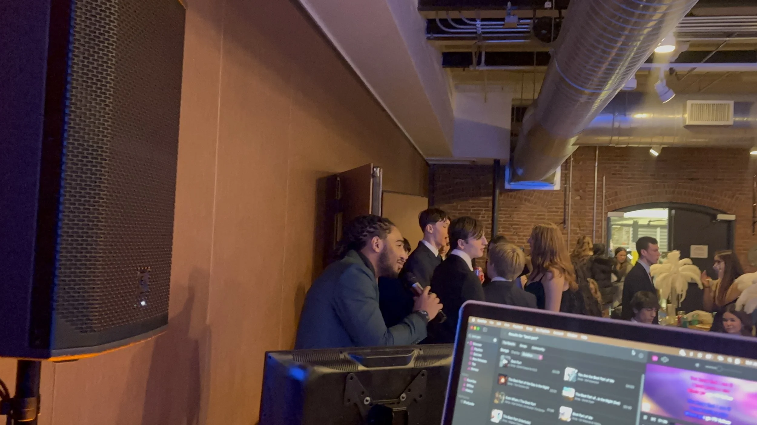 Young man holding microphone and speaking at a party or event with people standing in line and socializing inside a brick-walled venue.