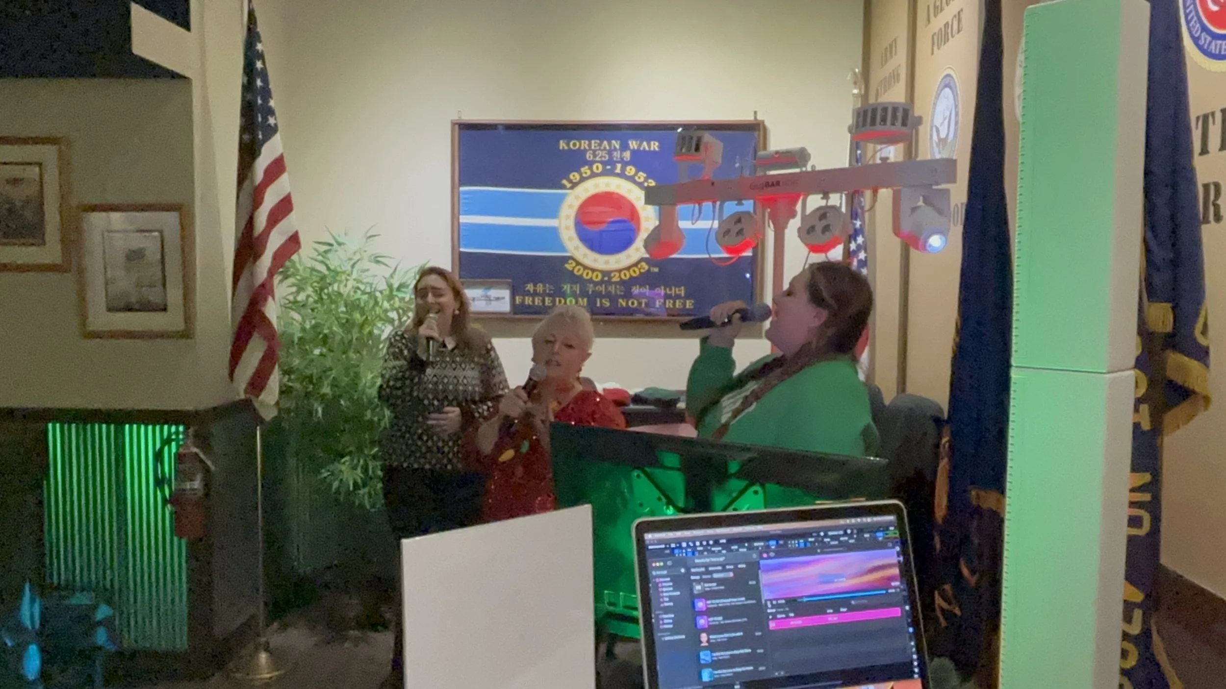 Three women singing karaoke in a room decorated with a Korean War memorial on the wall, an American flag, and a computer screen in the foreground.