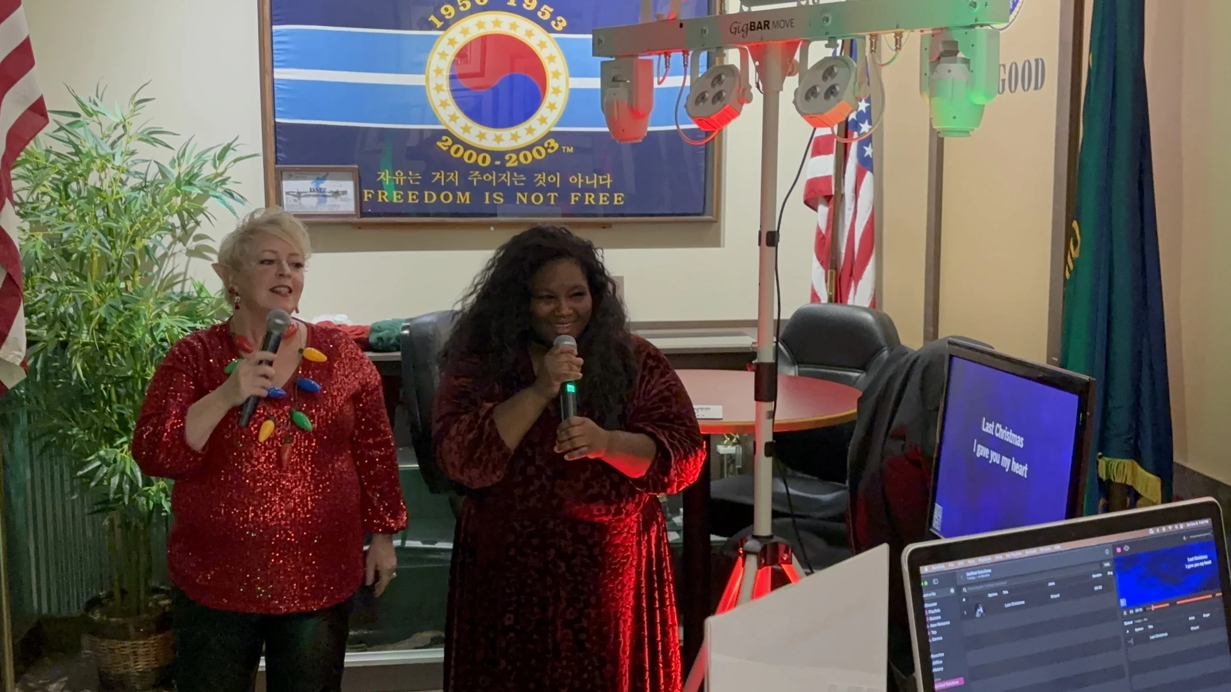 Two women singing with microphones in front of a presentation screen, decorated with Christmas lights and flags, in a room with a banner that reads "FREEDOM IS NOT FREE" and other text and symbols.