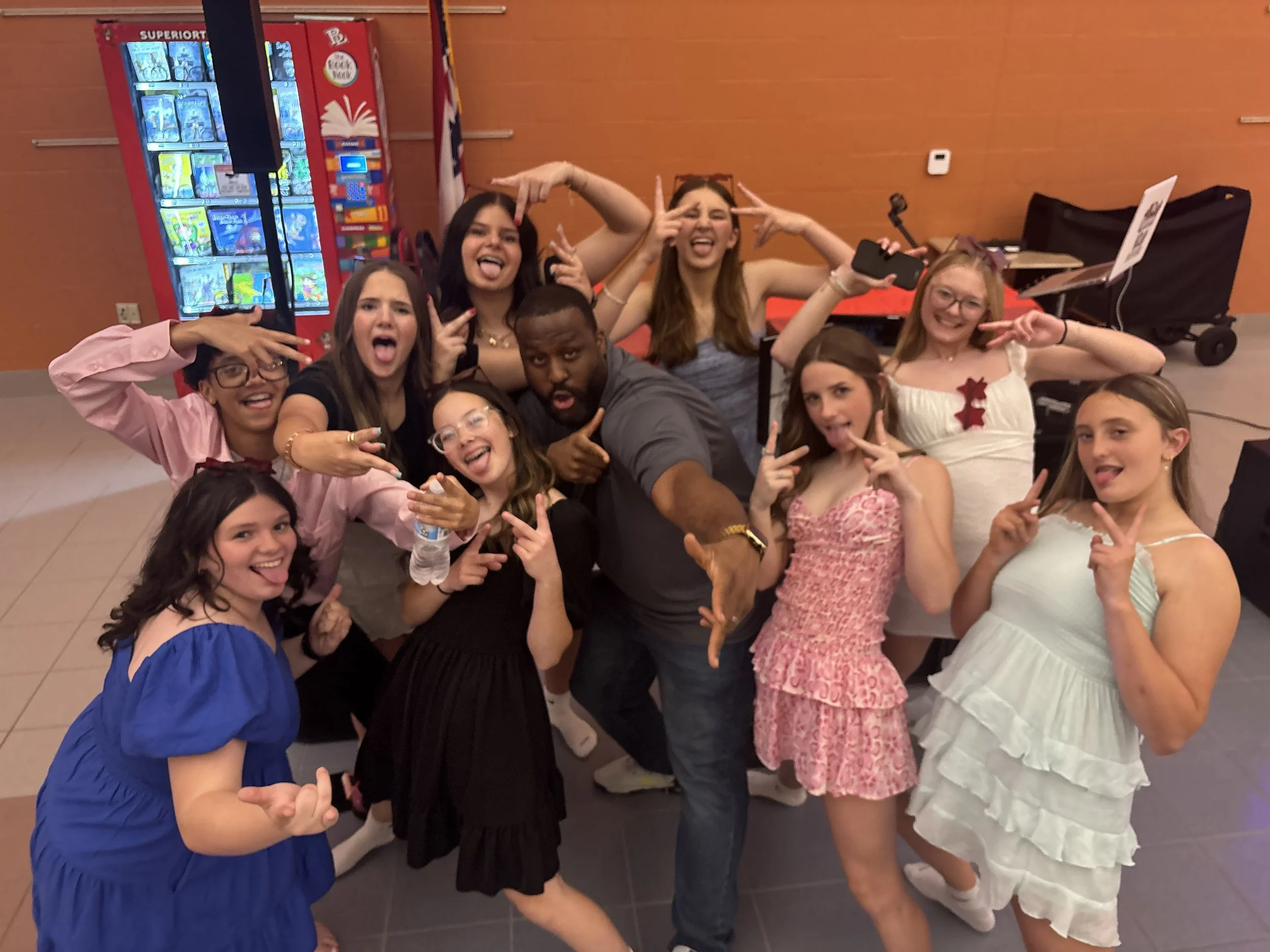 Group of young people and a man posing playfully in front of a vending machine and orange wall at indoor event